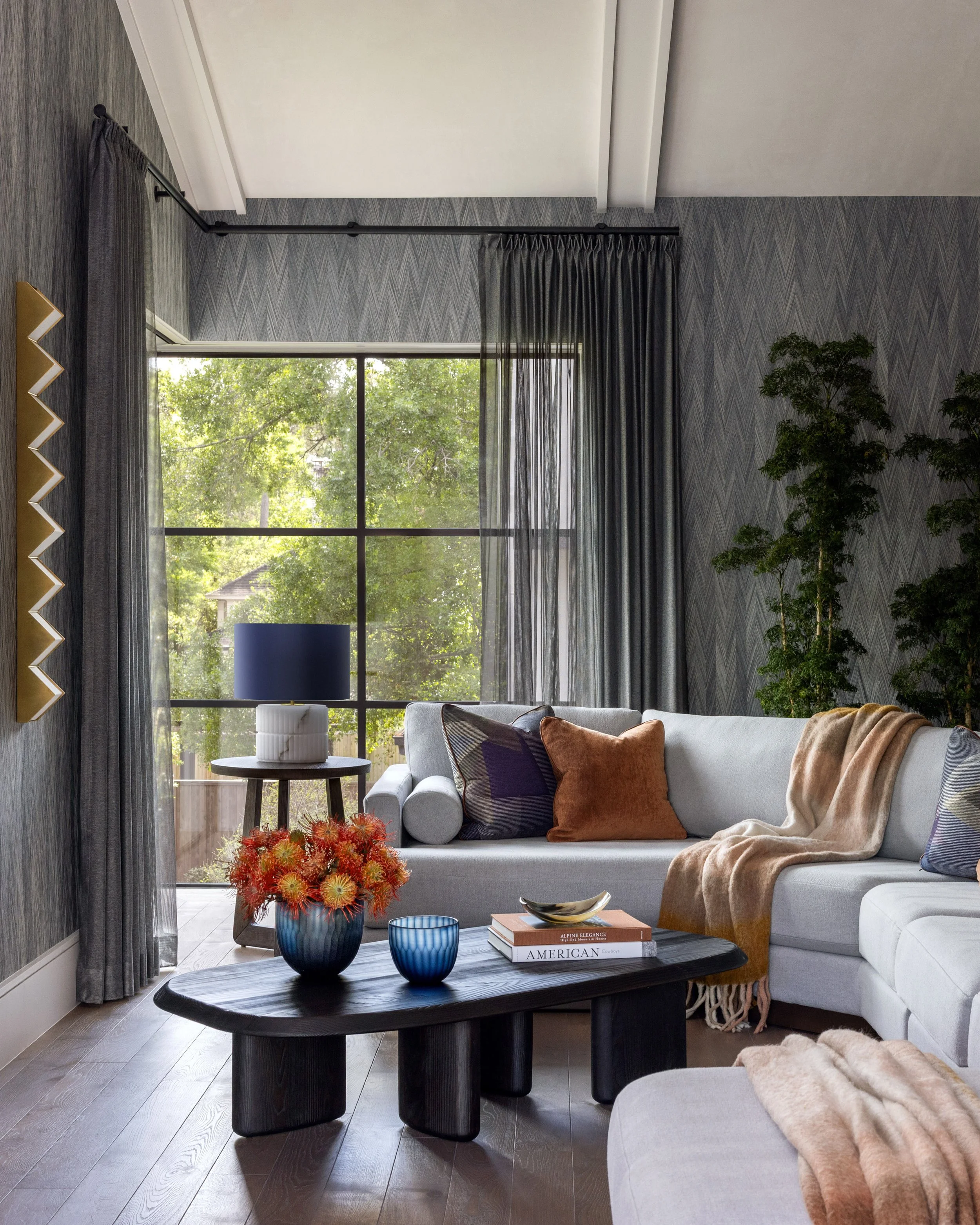 Benjamin Johnston Design living room featuring dark chevron grasscloth walls, gray sectional sofa with rust and purple accent pillows, sculptural black coffee table, floor-to-ceiling steel windows, sheer drapery, and orange floral arrangement.
