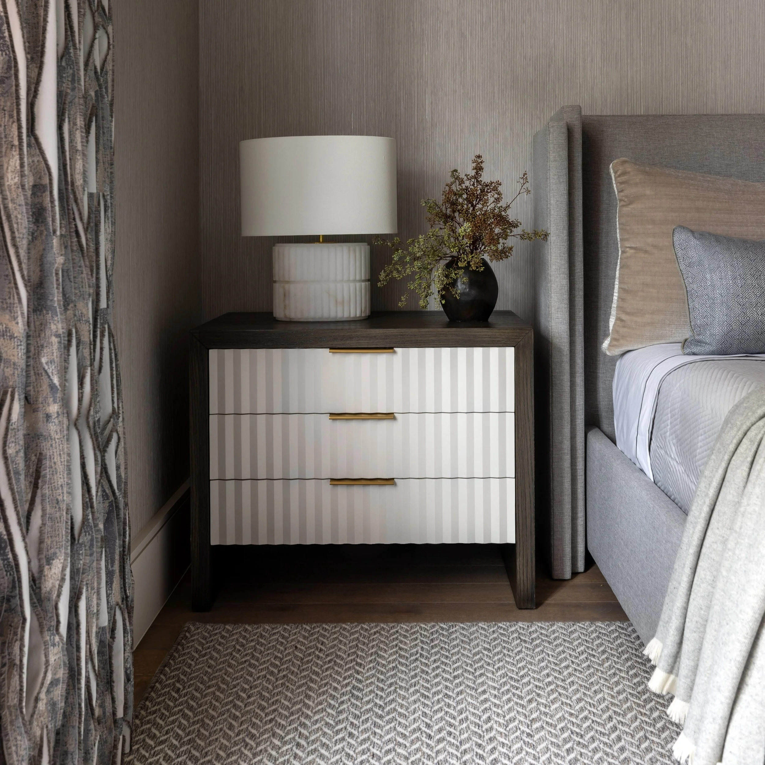 A bedside table with a white lamp and a vase with brown and green dried flowers, next to a bed with beige and gray pillows, in a room with textured beige wallpaper and patterned carpet.