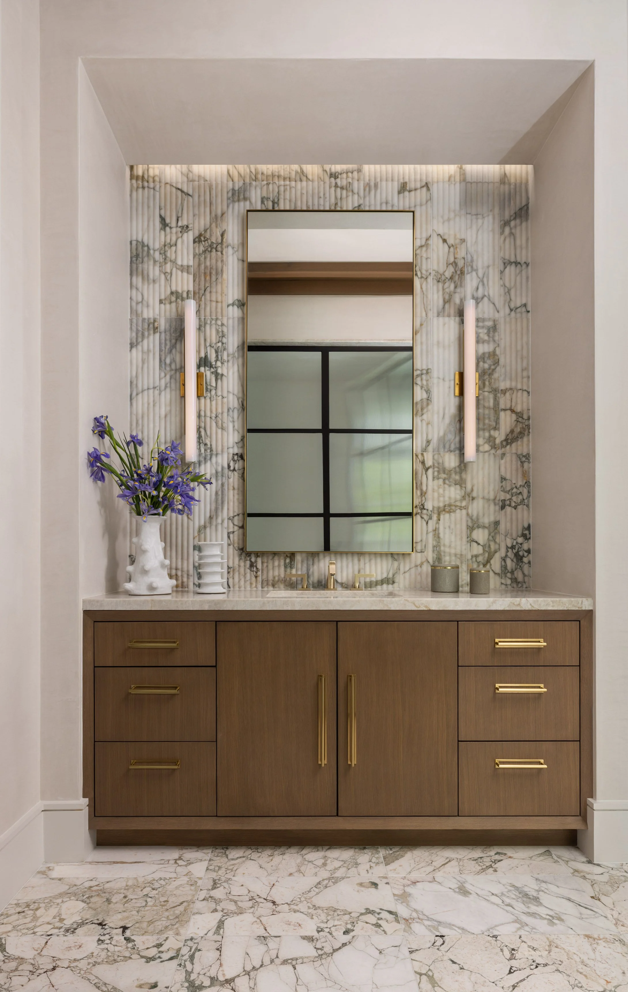 Bathroom vanity with wooden cabinet, marble countertop, wall mirror, wall-mounted lights, and decorative items including a vase with purple flowers and small containers, against marble wall and floor.