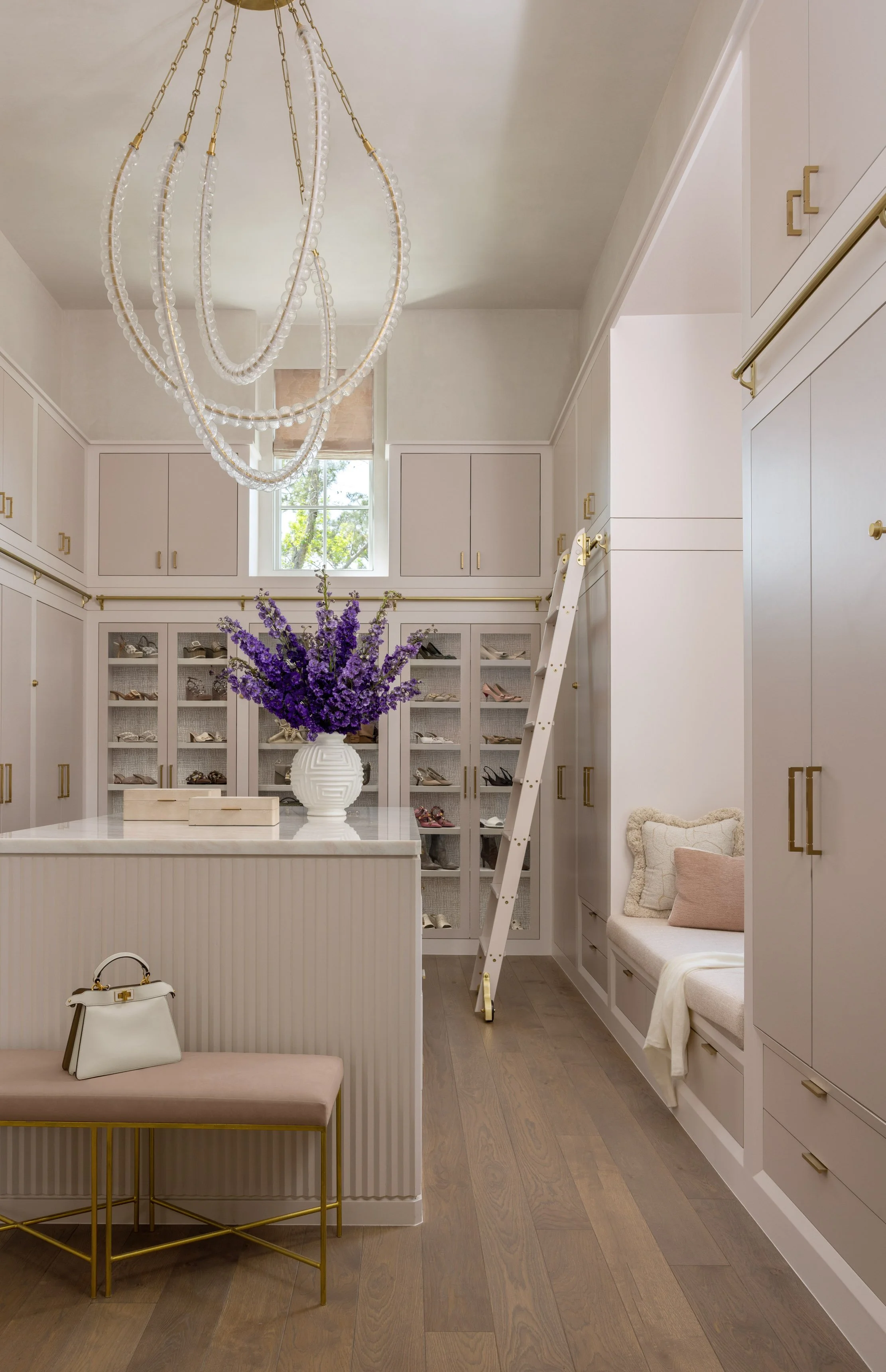 Interior view of a walk-in closet with white cabinetry, a large chandelier, a window, a purple flower arrangement, and a bench with a white handbag.