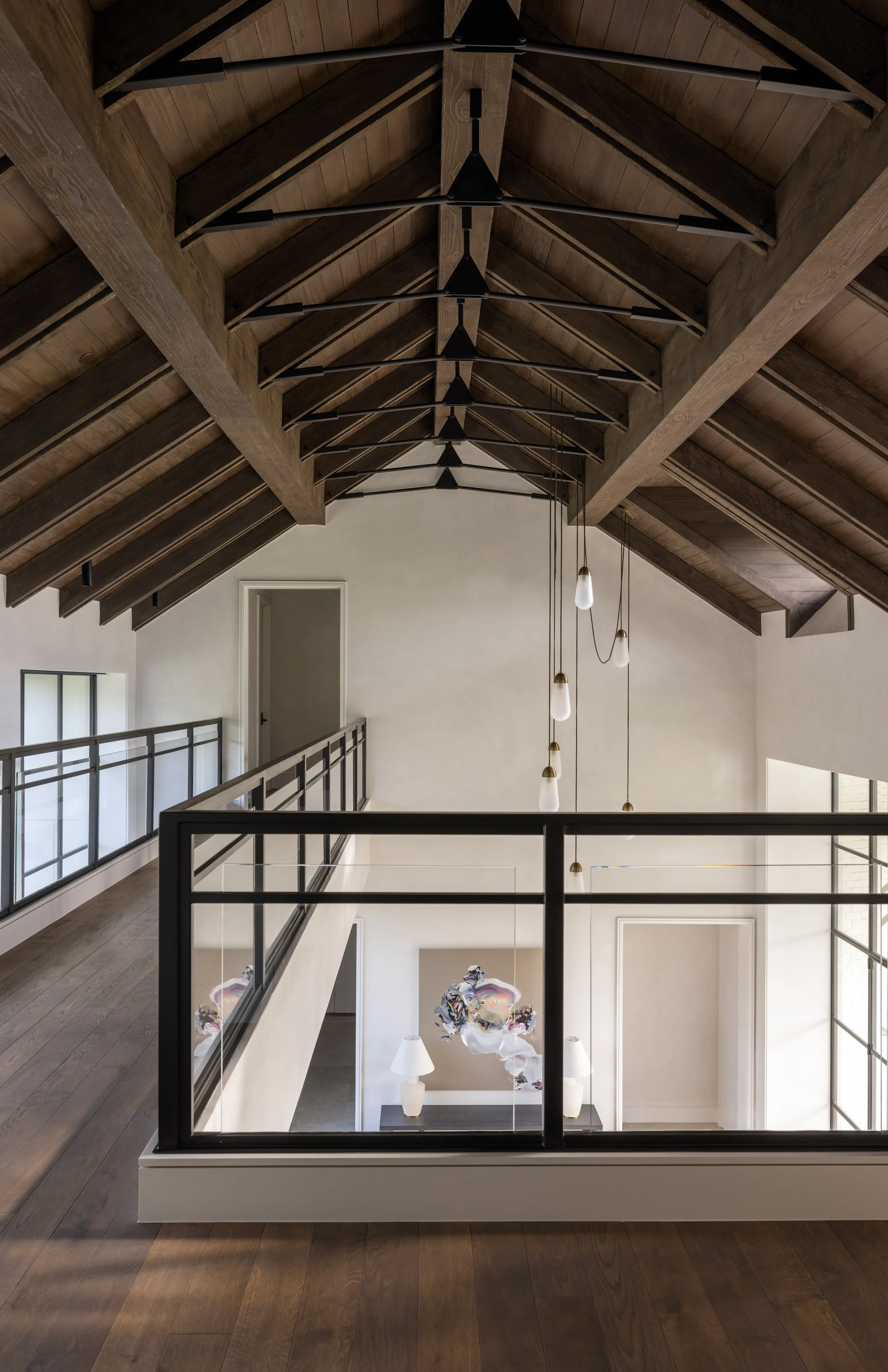 Benjamin Johnston Design upper hallway featuring soaring vaulted ceiling with exposed wood trusses, black steel railing, cascading pendant lights, and view into entryway below with abstract artwork.