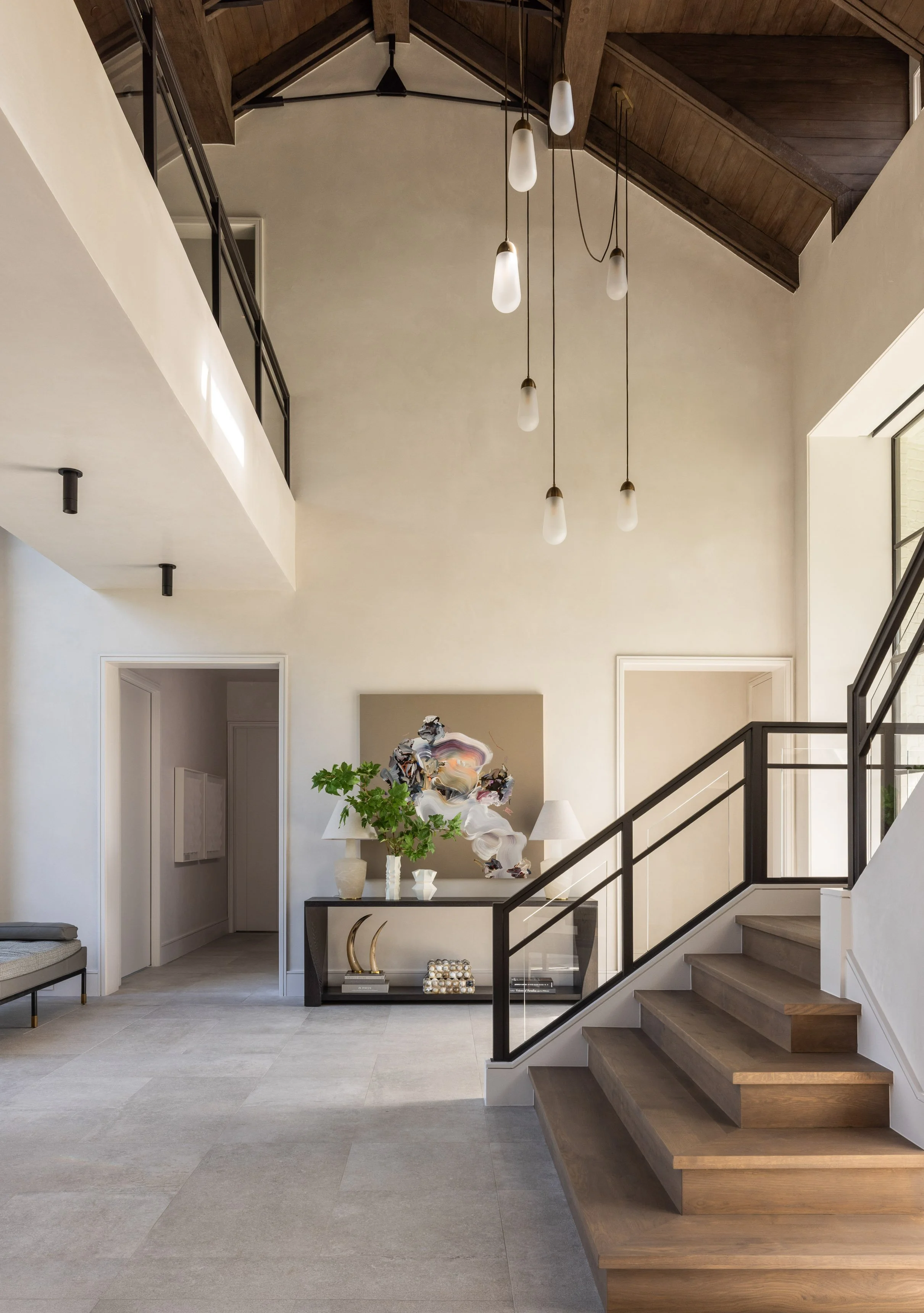 Benjamin Johnston Design entryway featuring soaring vaulted ceiling with exposed wood trusses, cascading pendant lights, black steel staircase railing, dark console table with abstract artwork, and stone tile floors.