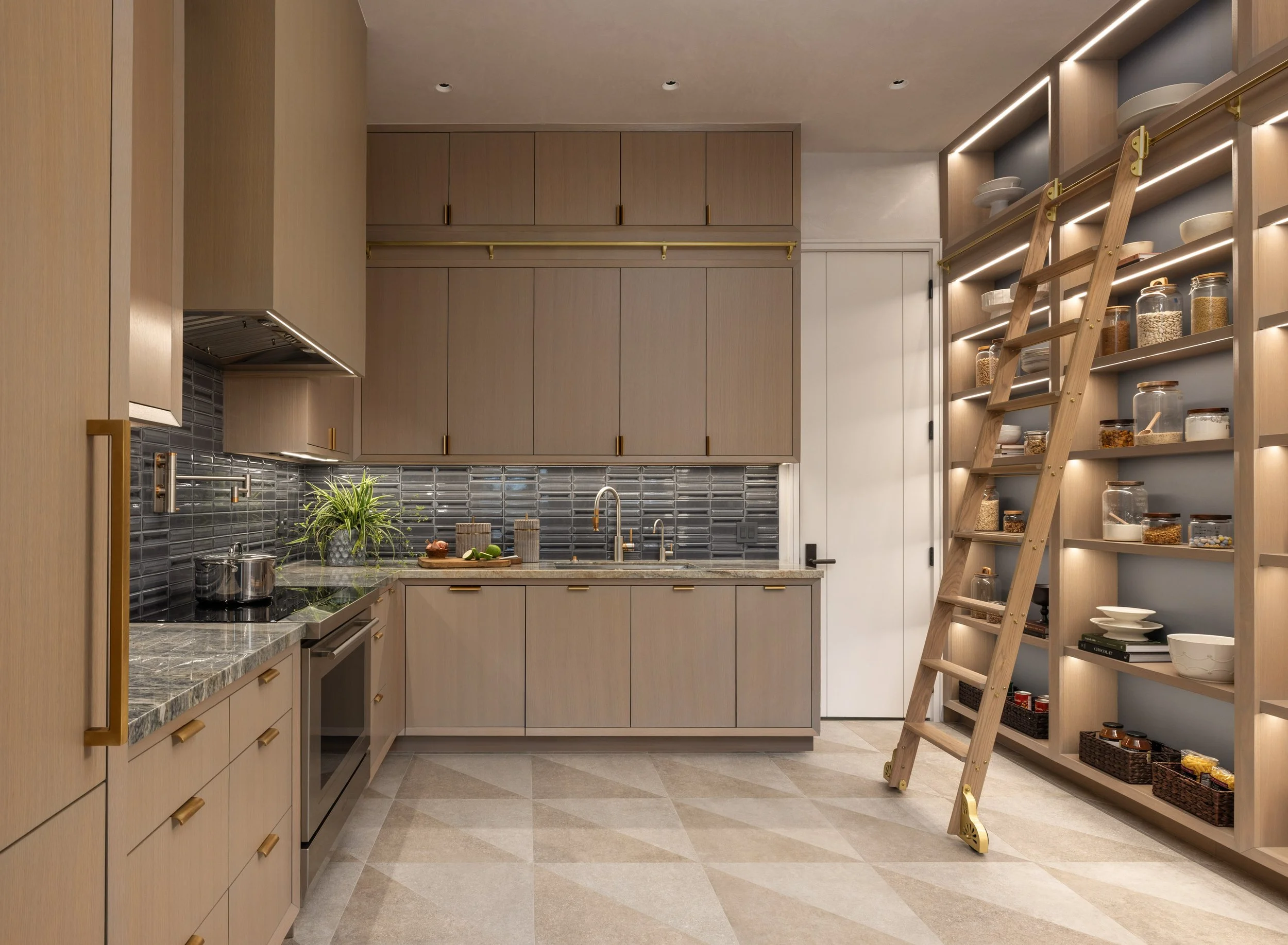 Benjamin Johnston Design Houston catering kitchen featuring taupe flat front cabinetry with brass hardware, dark glazed tile backsplash, marble countertops, geometric stone tile floor, and illuminated floor-to-ceiling pantry shelving with brass.