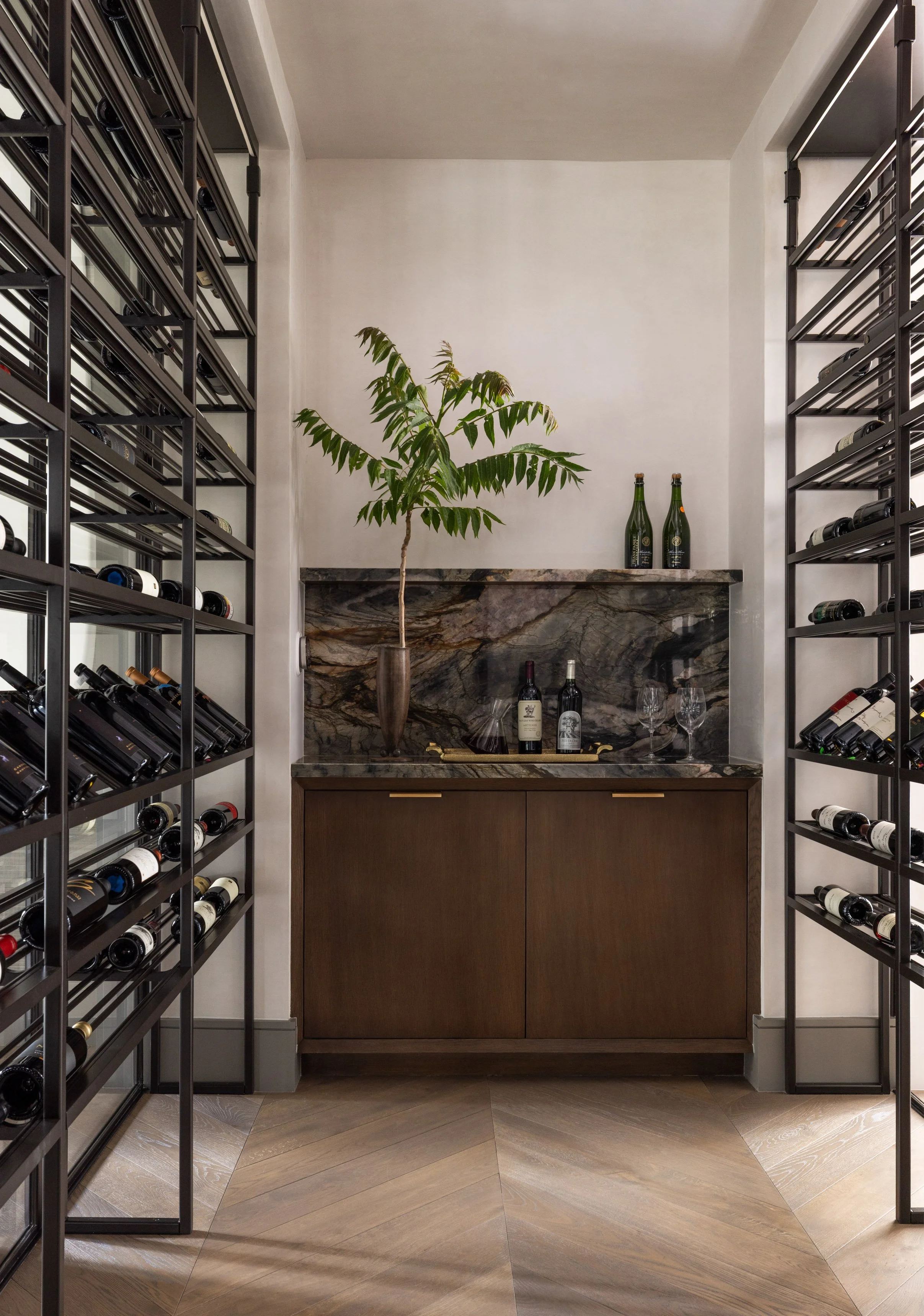 Benjamin Johnston Design wine room with floor to ceiling dark metal wine racks, walnut cabinet with brass hardware, veined marble backsplash and countertop, potted plant, wine bottles, and herringbone wood floor.