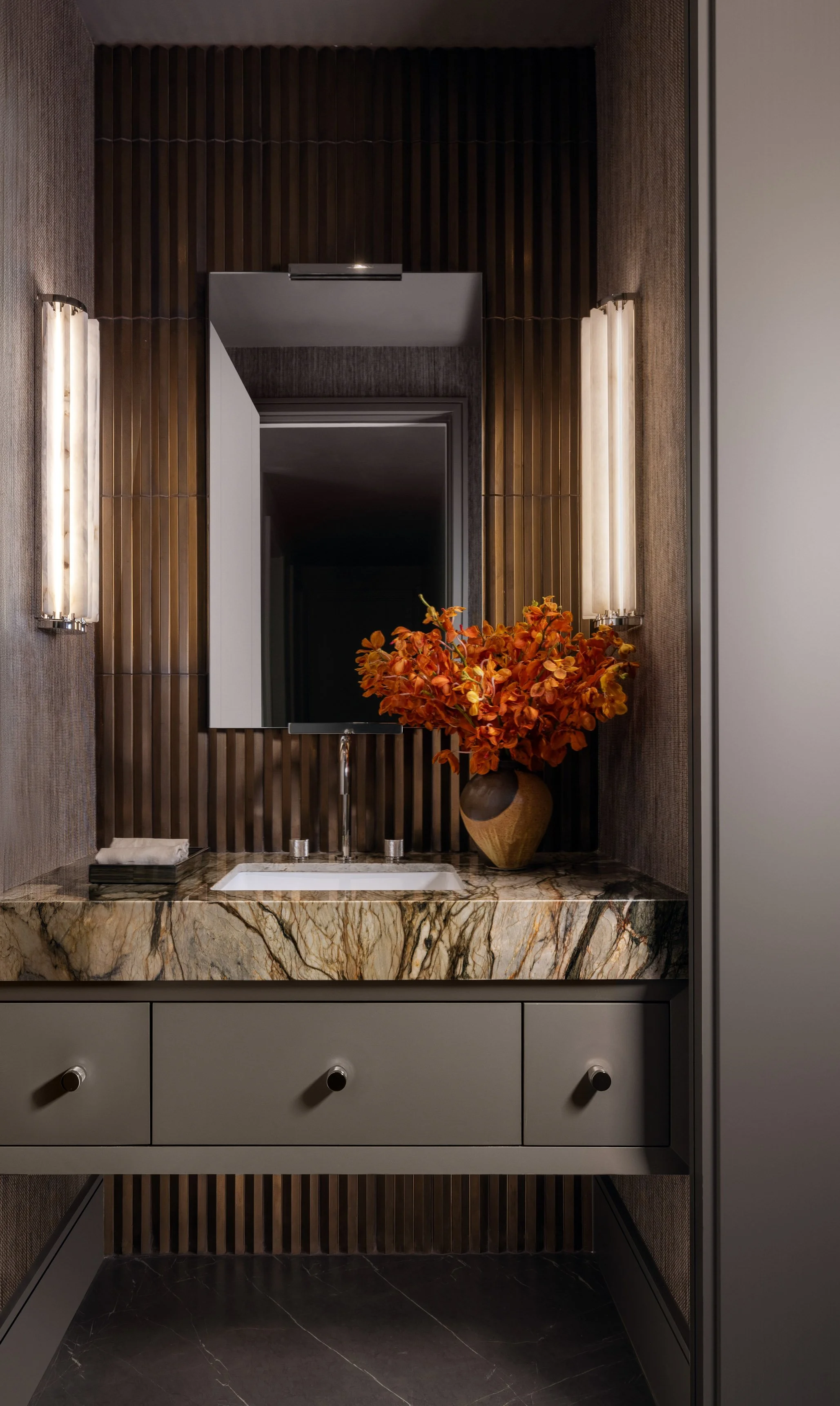 A modern bathroom vanity with a marble countertop, a sink with a chrome faucet, and a large mirror. The vanity has three drawers with black knobs. A large vase with orange and red flowers is placed on the countertop. The wall behind the mirror featur