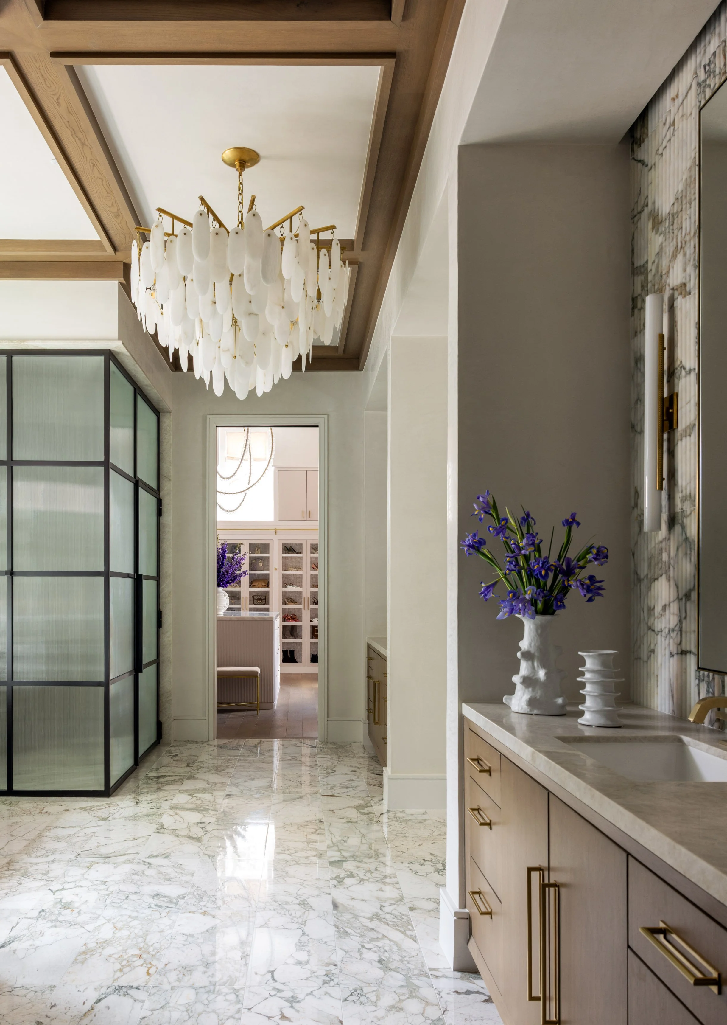 Interior of a modern home with a marble floor, a wooden console with a white vase of purple flowers, and a large white chandelier with gold accents hanging from the ceiling.