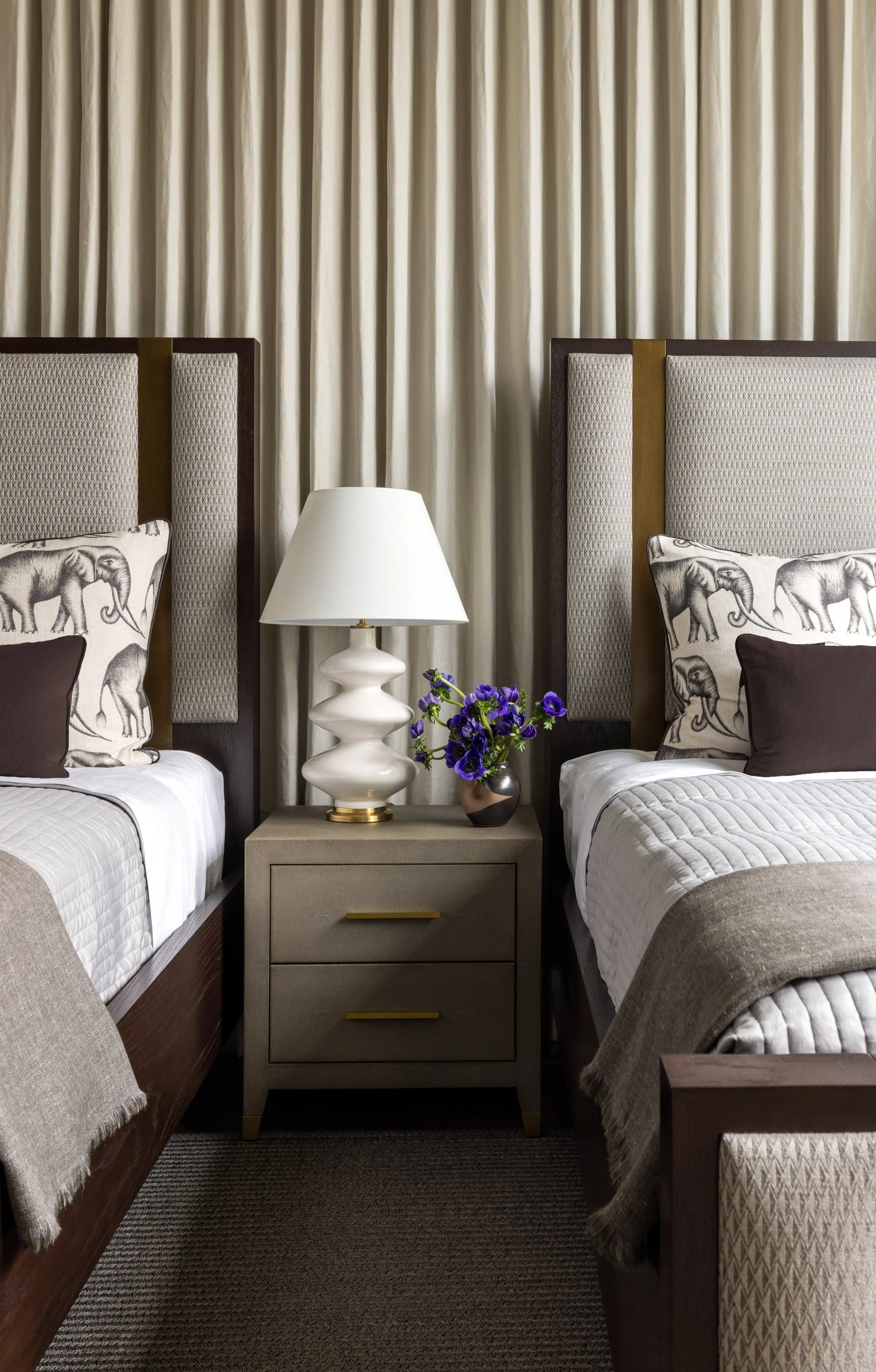 Two beds with elephant-patterned pillows in a hotel room, a nightstand with a white lamp and purple flowers, beige curtains behind the beds.