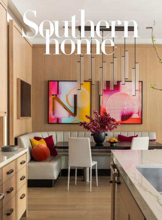 Modern dining area with a built-in white sectional sofa, colorful throw pillows, a black dining table with two white chairs, a large vase of pink flowers, abstract wall art, a unique pendant light fixture, and a kitchen countertop in the foreground.
