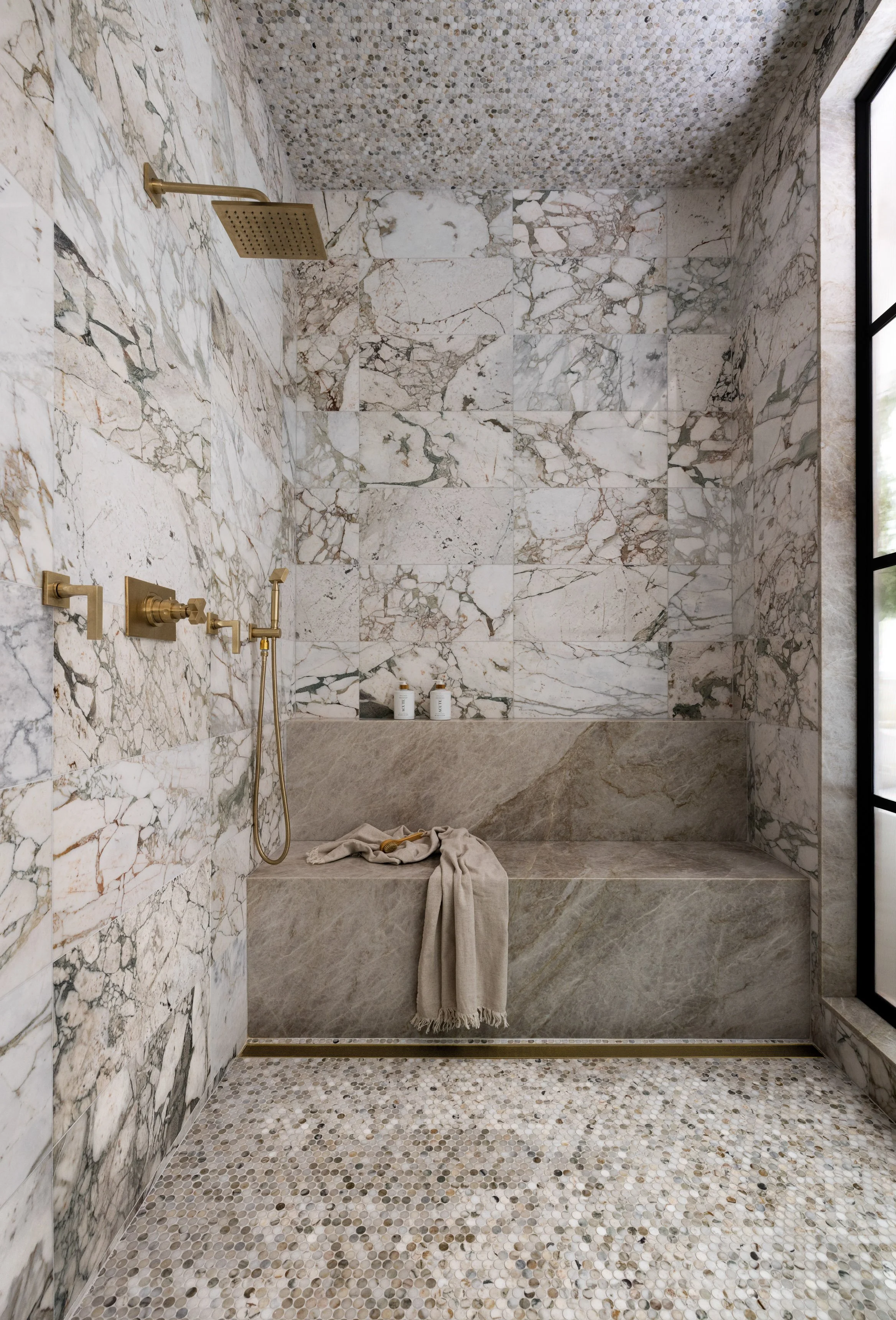 A walk-in shower with marble walls, a built-in bench, a rainfall showerhead, and a handheld shower with gold fixtures. The floor is covered in small, round mosaic tiles, and a beige towel hangs over the bench.