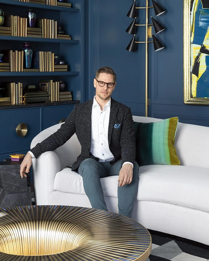 A man in glasses and a dark blazer sitting on a white couch in a modern, colorful living room with blue walls, bookshelves, and a large abstract painting.