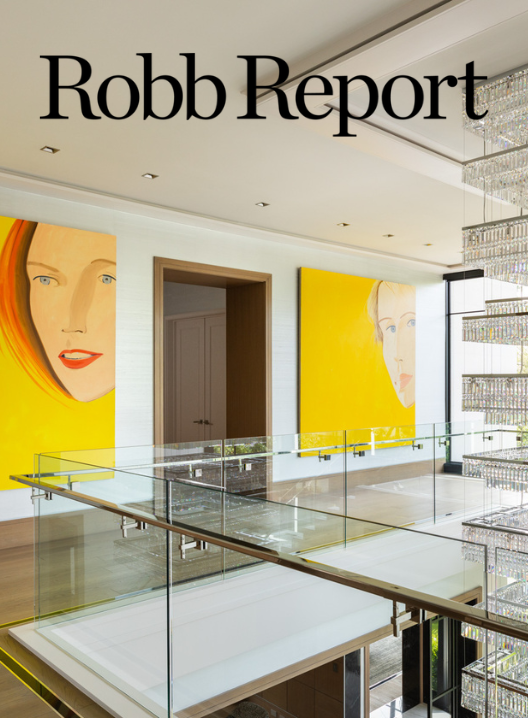Benjamin Johnston Design featured in Robb Report May 2026, showcasing a luxury Houston home staircase landing with glass and brass railing, oversized bold yellow figurative art panels, and a cascading crystal chandelier.