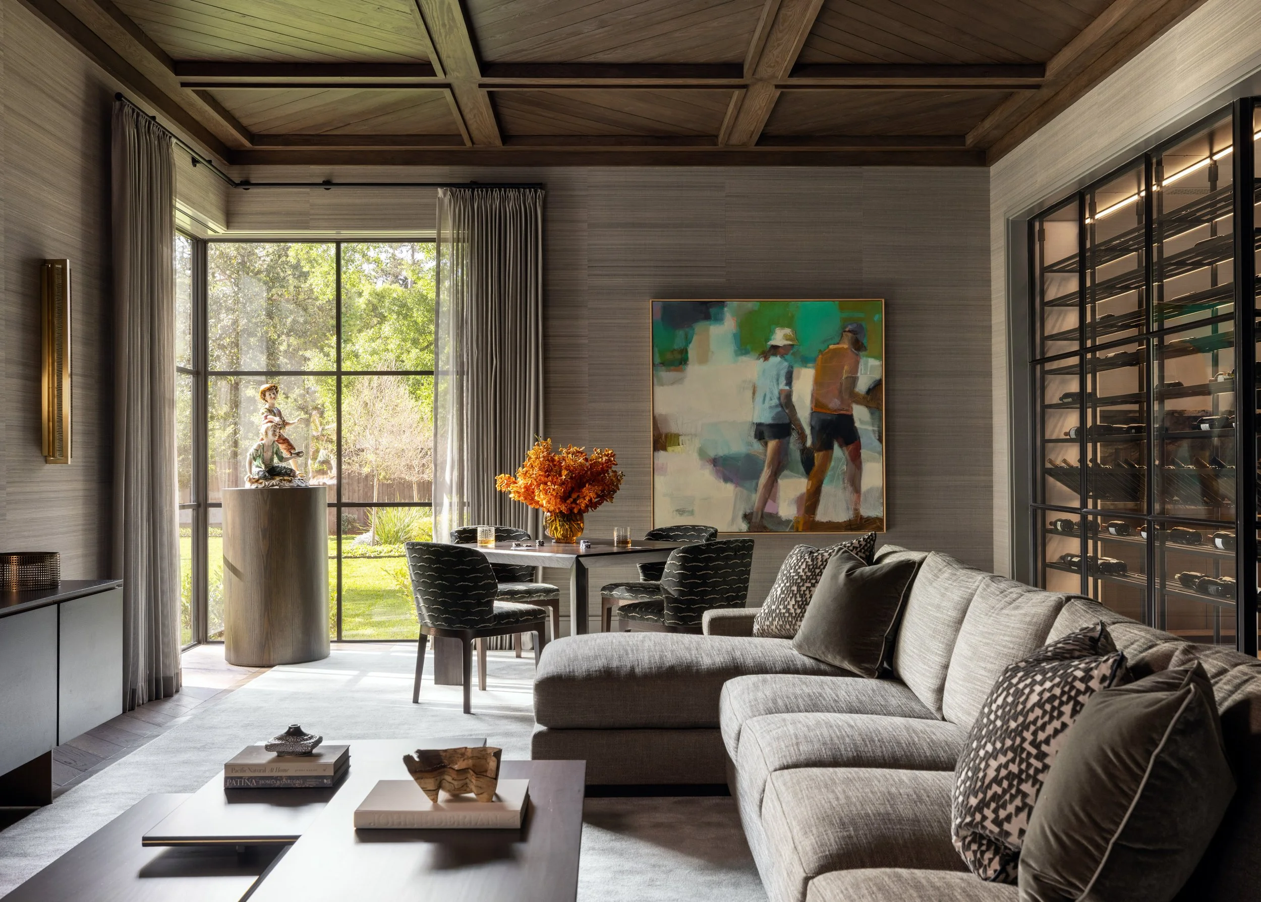 Modern living room with large window, artwork on the wall, gray sofa with patterned pillows, black dining table with chairs, and a coffee table with books and decorative items.