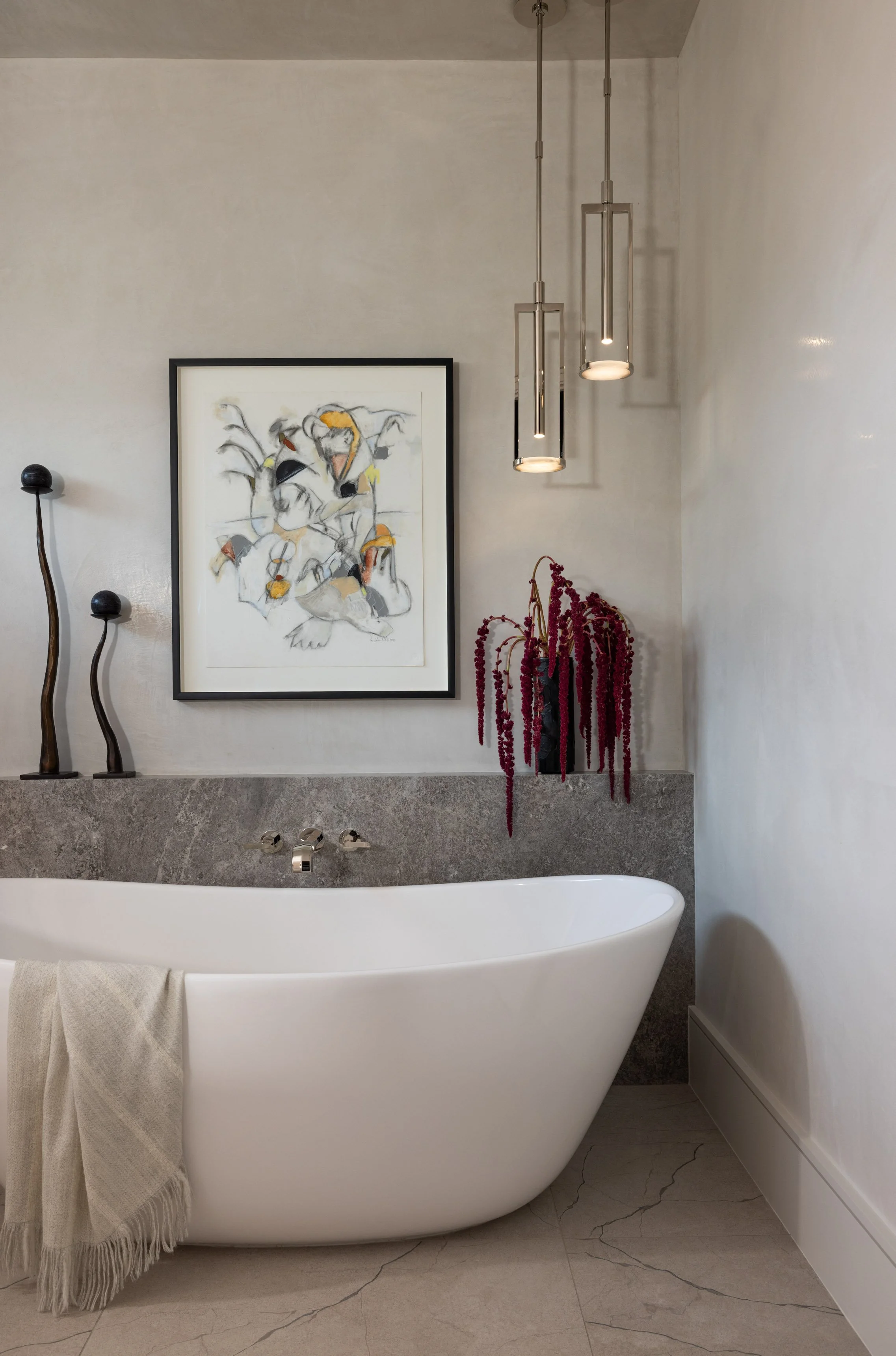A modern bathroom featuring a white bathtub, a grey stone backsplash, black decorative sculptures, a framed artwork of a bird, and pendant lights hanging from the ceiling. There is a towel hanging on the edge of the bathtub.