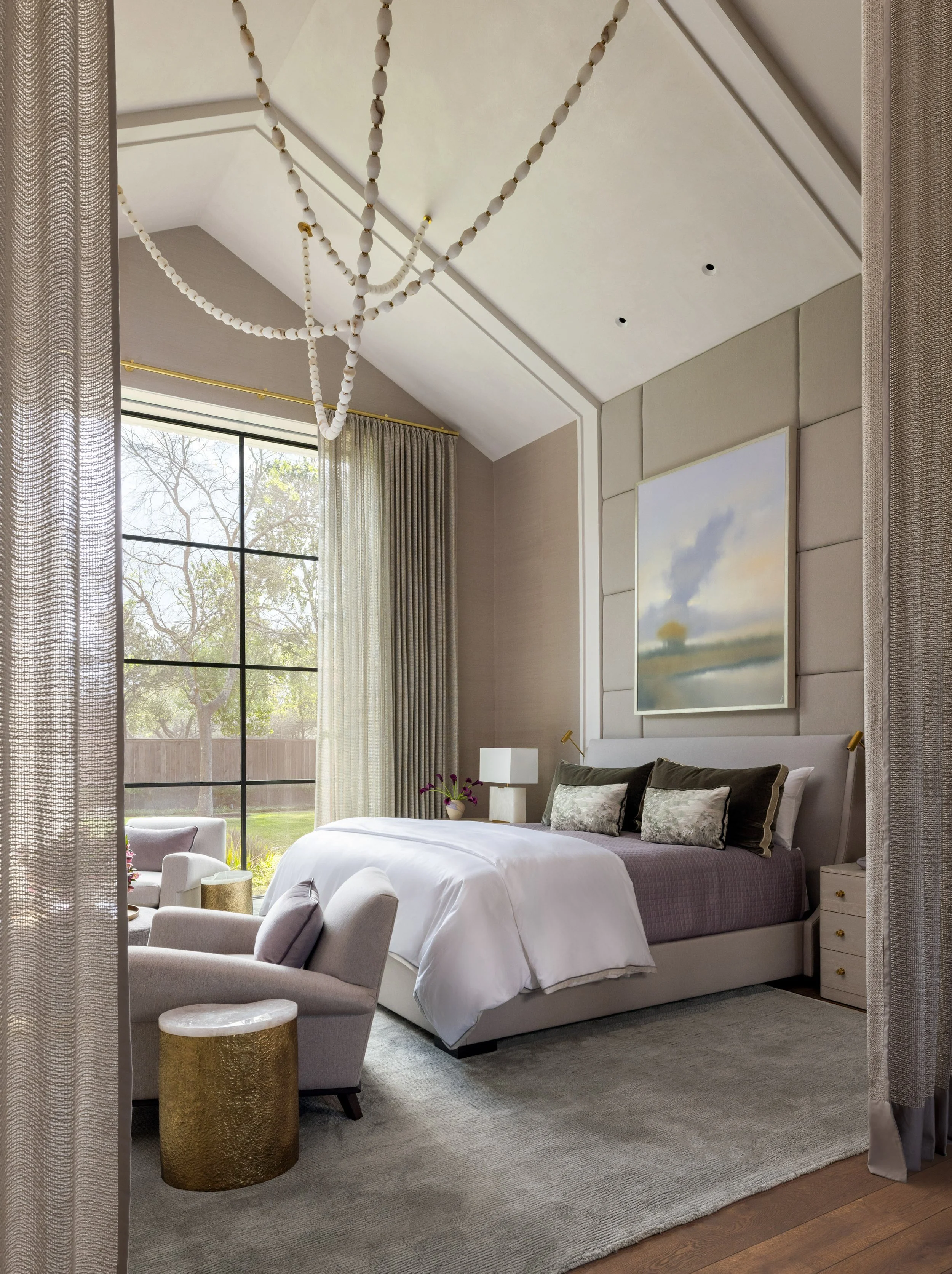 Benjamin Johnston Design Houston primary bedroom featuring sculptural beaded chandelier, upholstered panel headboard wall, soft landscape artwork, vaulted ceiling, floor-to-ceiling steel windows, and brass accent side table.