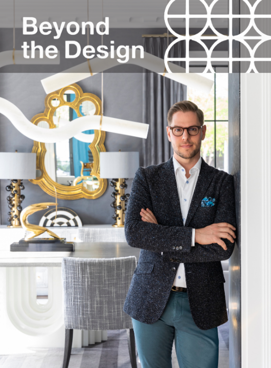 A man standing with arms crossed in a modern, stylish interior room featuring a large gold mirror, a table lamp, and a patterned wall decoration.