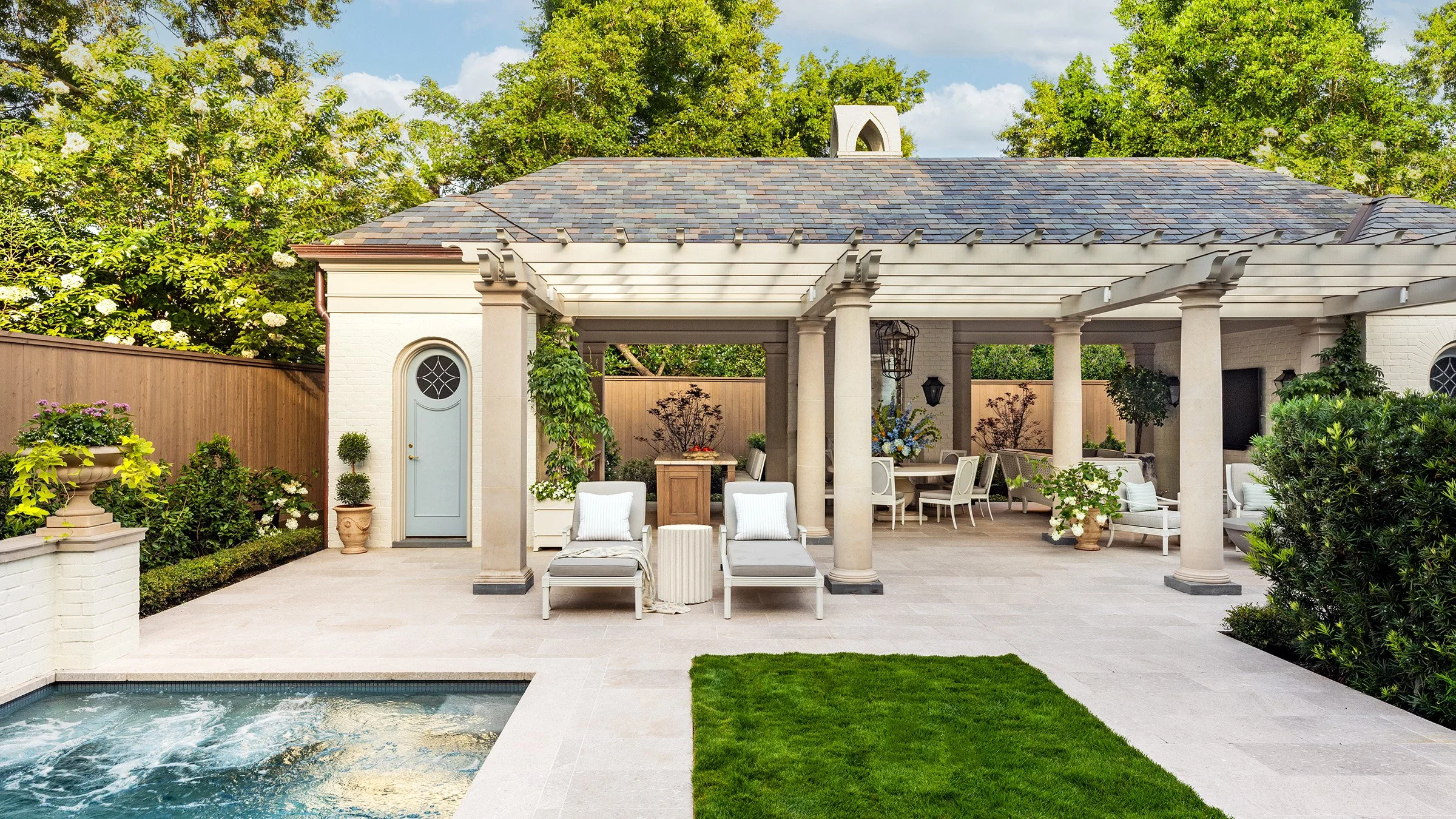 A luxurious outdoor living pavilion featuring classical stone columns, a covered terrace with elegant lounge and dining areas, a slate roof, limestone flooring, and a relaxing spa. Surrounded by manicured gardens, mature trees, and tailored landscapi