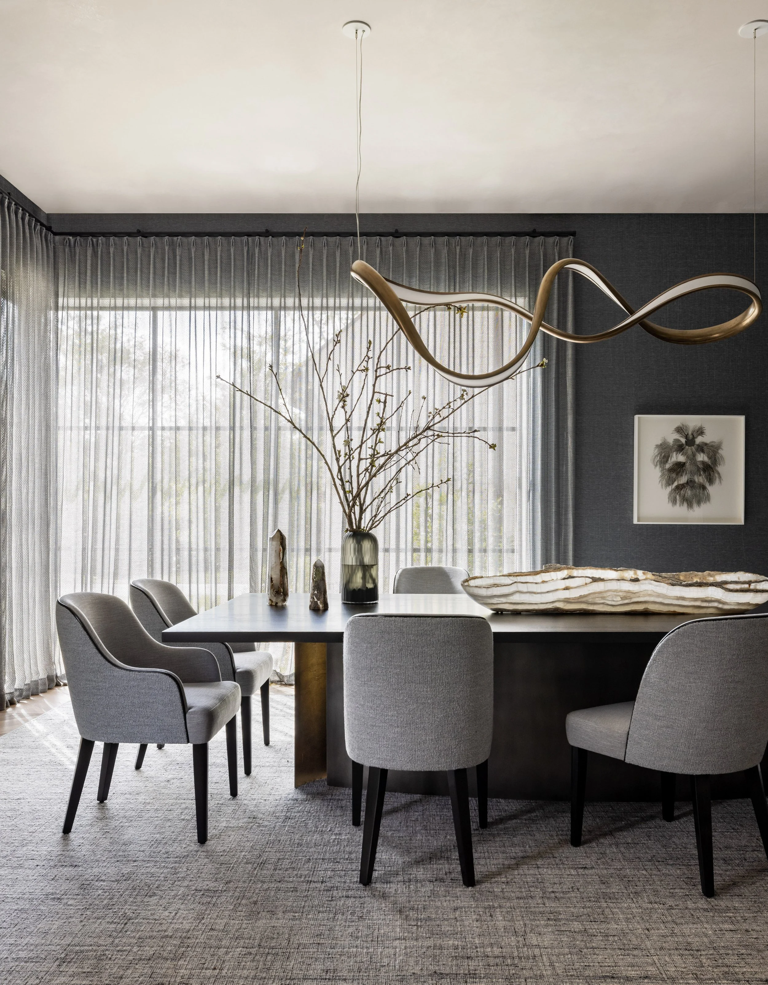 Modern dining room with gray upholstered chairs, a black dining table, decorative vases, a large shell-shaped centerpiece, a glass vase with branches, and a framed art print of a plant on a dark gray wall, with floor-to-ceiling sheer curtains allowin