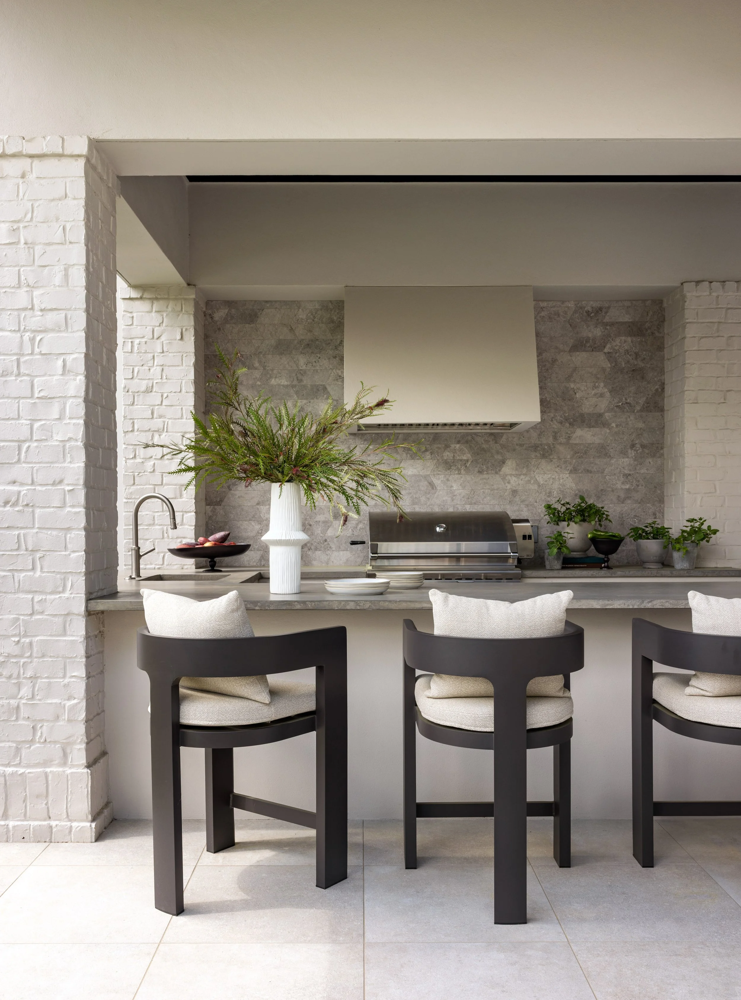 Benjamin Johnston Design Houston outdoor kitchen featuring stone tile backsplash, concrete countertop, built-in grill, white range hood, white brick surround, and dark wood bar stools with linen cushions.