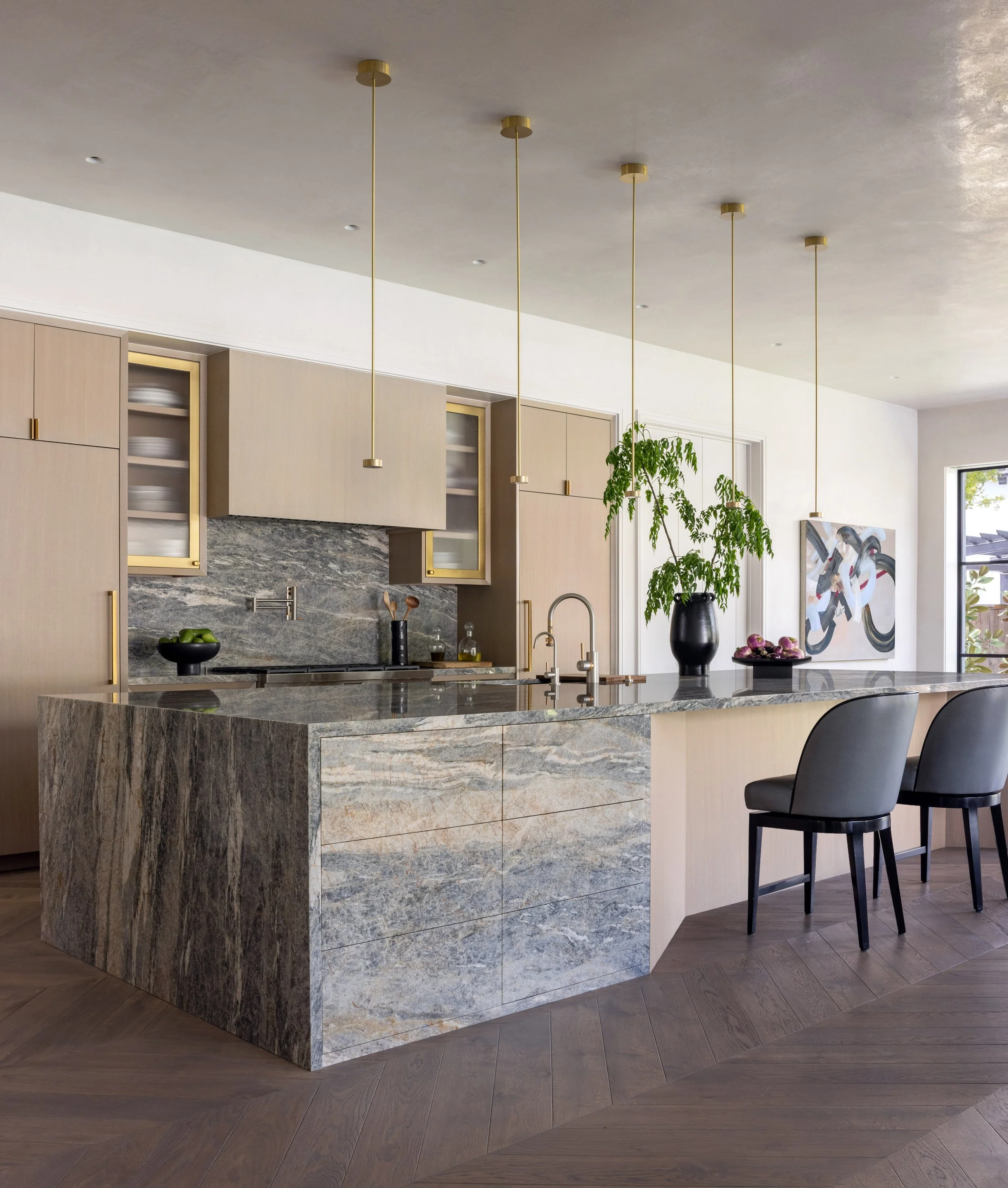 Benjamin Johnston Design kitchen featuring dramatic marble waterfall island, brass pendant lights, flat front cabinetry with brass framed glass uppers, veined marble backsplash, gray leather bar stools, herringbone wood floor, and abstract artwork.