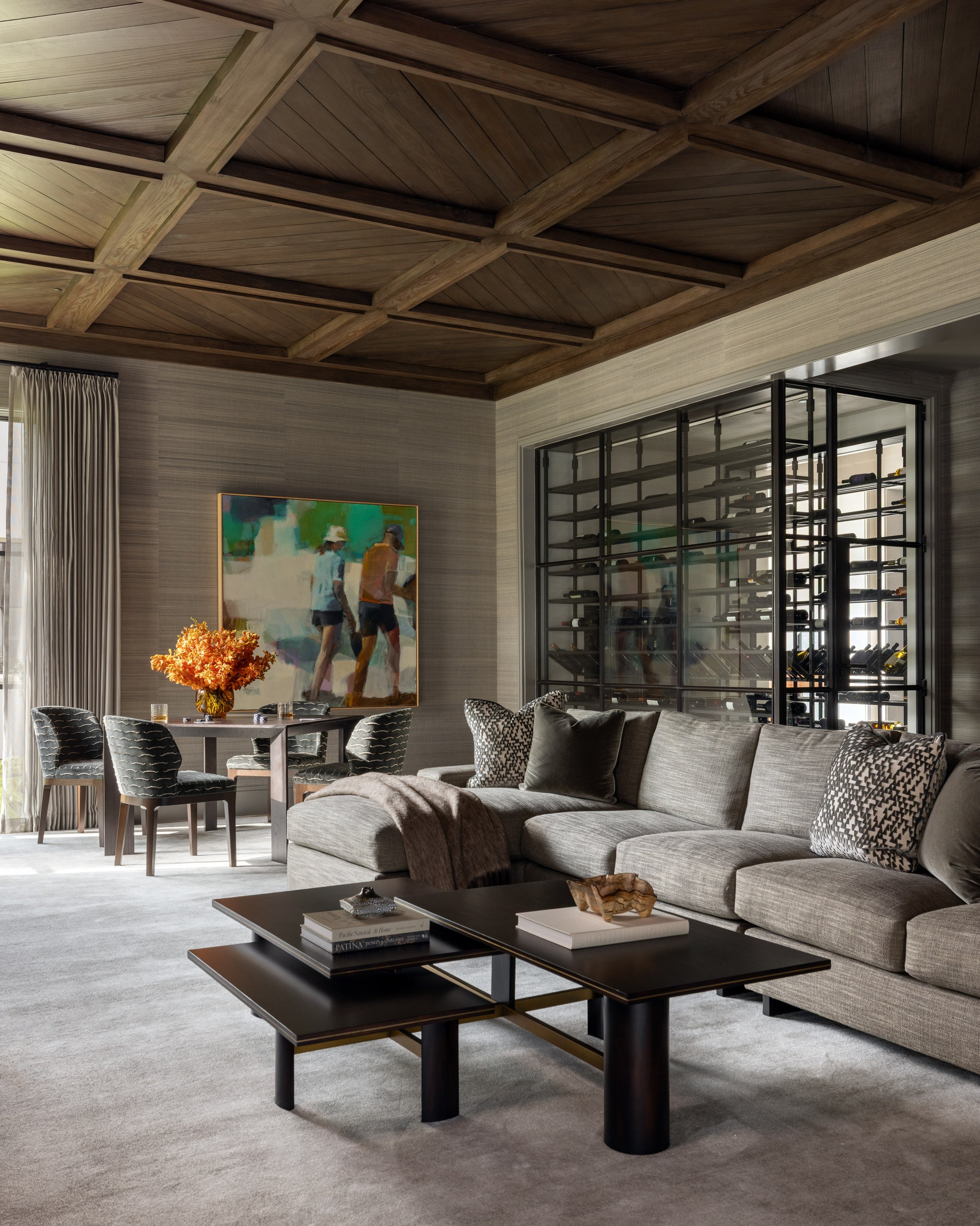 Modern living room with a gray sofa, patterned throw pillows, a black coffee table with books and decorative items, a dining table with black chairs, an abstract painting on the wall, a large window with beige curtains, and a wood-paneled coffered ce