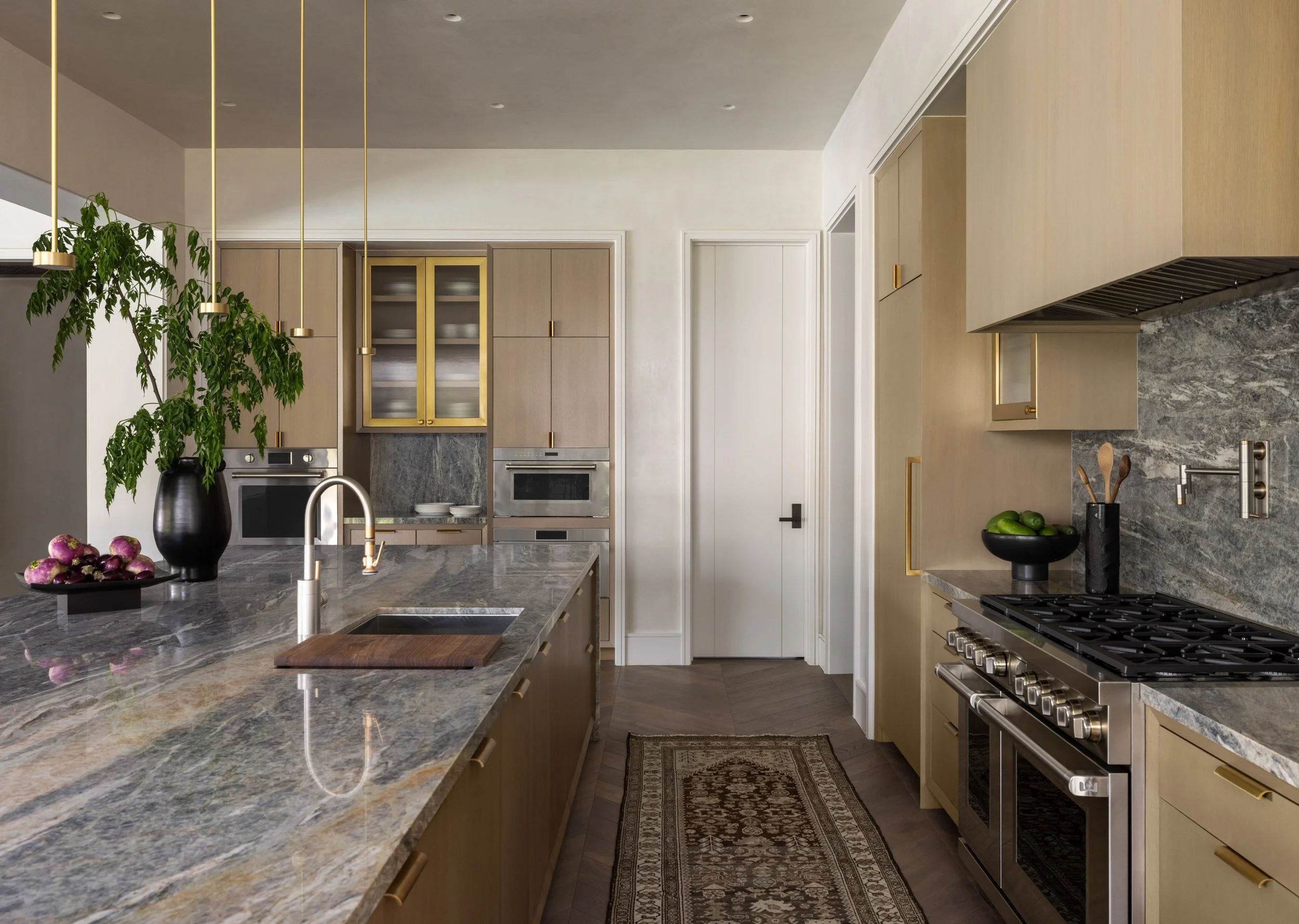 A modern kitchen with a large marble countertop island, light wood cabinets, stainless steel oven and range, and decorative details like a potted plant and bowls of fruit.