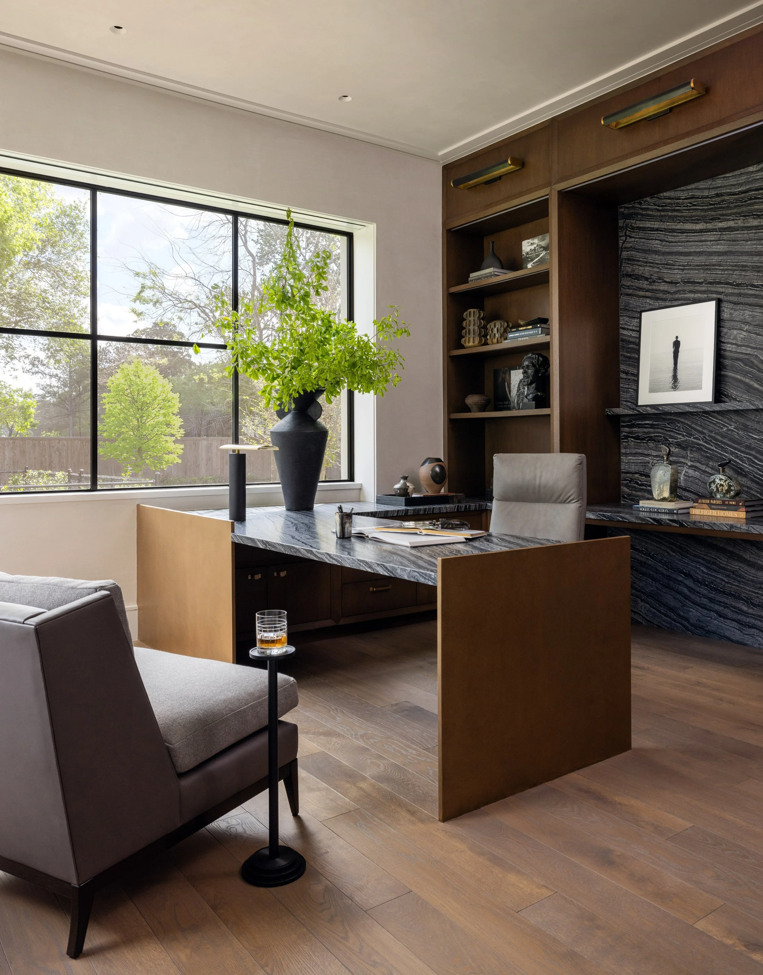 Benjamin Johnston Design home office with marble topped brass desk, dark stone accent wall, walnut built-in shelving with brass picture lights, gray lounge chair, black ceramic vase with greenery, and wide plank wood floor.