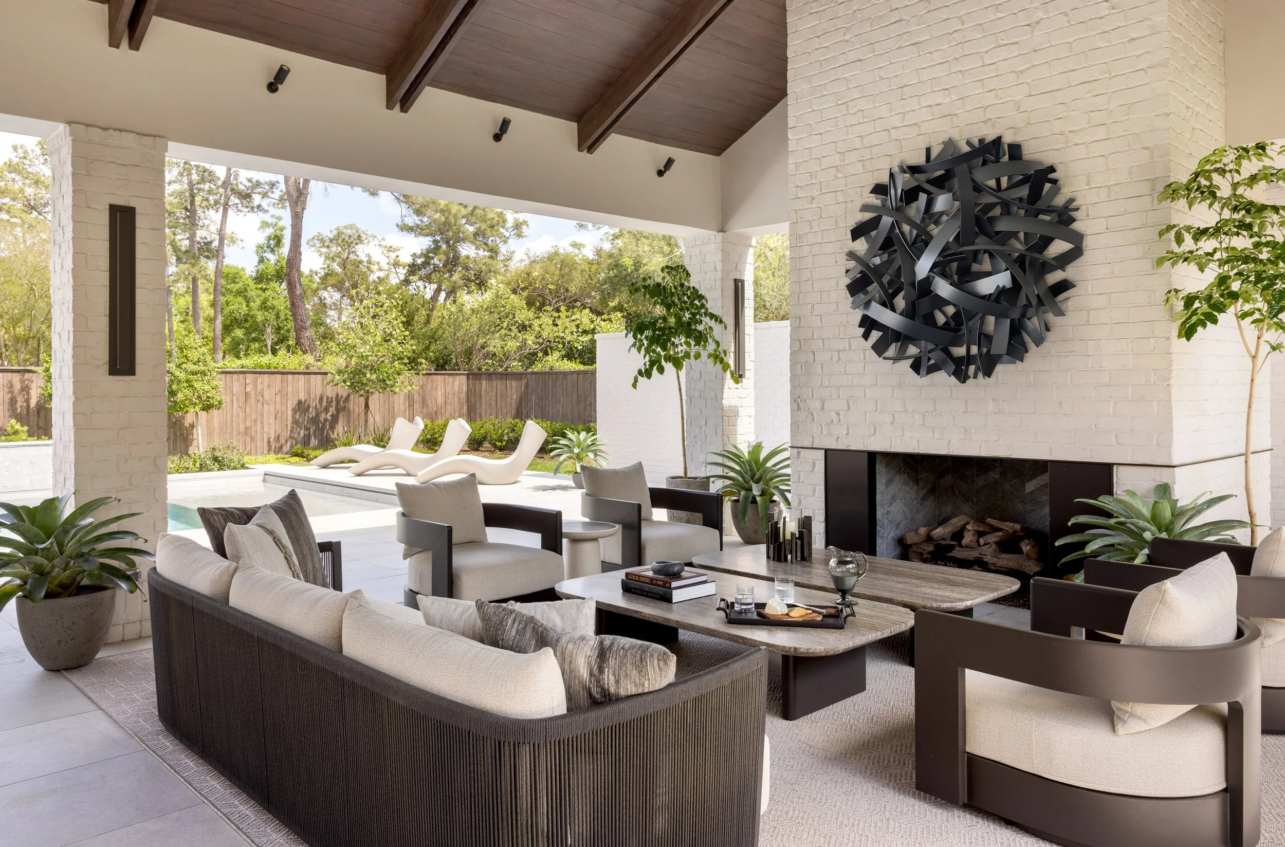 Benjamin Johnston Design outdoor lounge with vaulted wood beam ceiling, woven sofa and chairs with linen cushions, stone coffee table, white brick fireplace, large abstract metal wall sculpture, and pool view beyond.