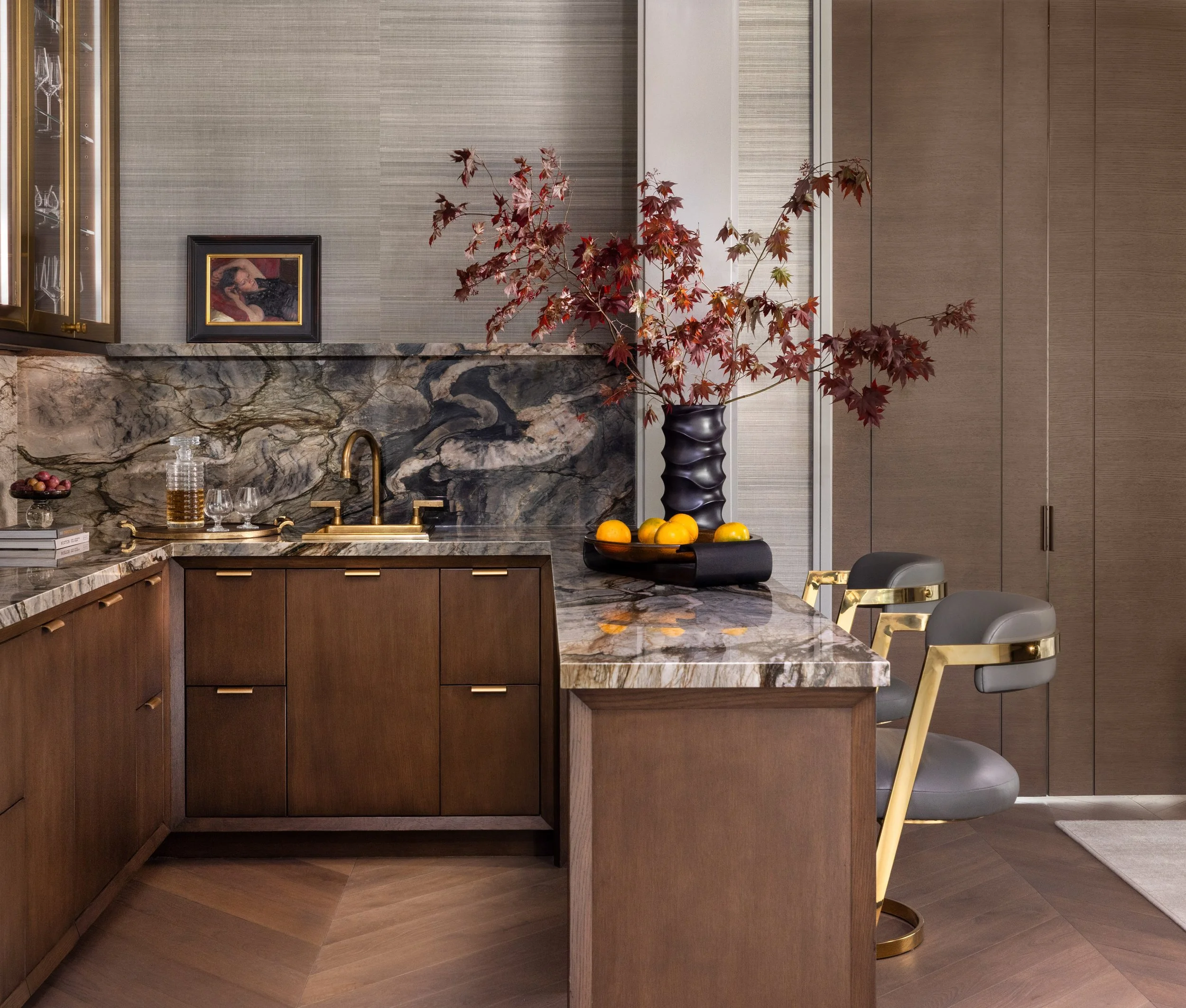 Benjamin Johnston Design wet bar with book matched marble backsplash and island countertop, dark walnut cabinetry with brass hardware, brass faucet, sculptural bar stool with brass frame, red maple branches, and herringbone wood floor.