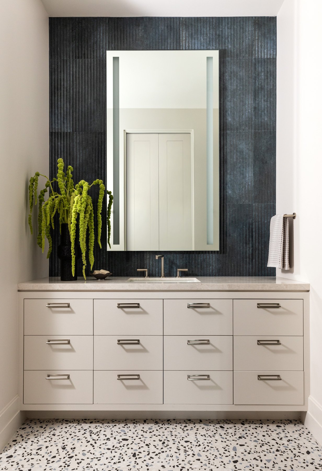 Benjamin Johnston Design Houston luxury residential interior design — modern bathroom vanity with custom cream cabinetry, marble countertop, backlit LED mirror, dark textured tile accent wall, terrazzo floor tile, and brushed nickel hardware.