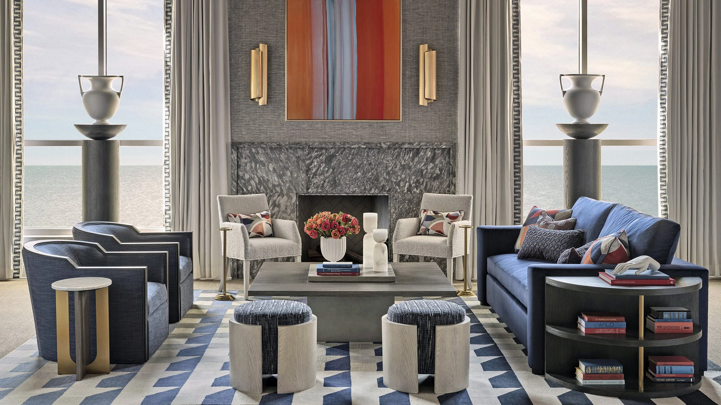 Living room with a fireplace, two white armchairs, a blue couch, two framed armchairs, a coffee table with a vase of pink flowers and decorative objects, large windows overlooking the ocean, abstract artwork, and various books and decor.