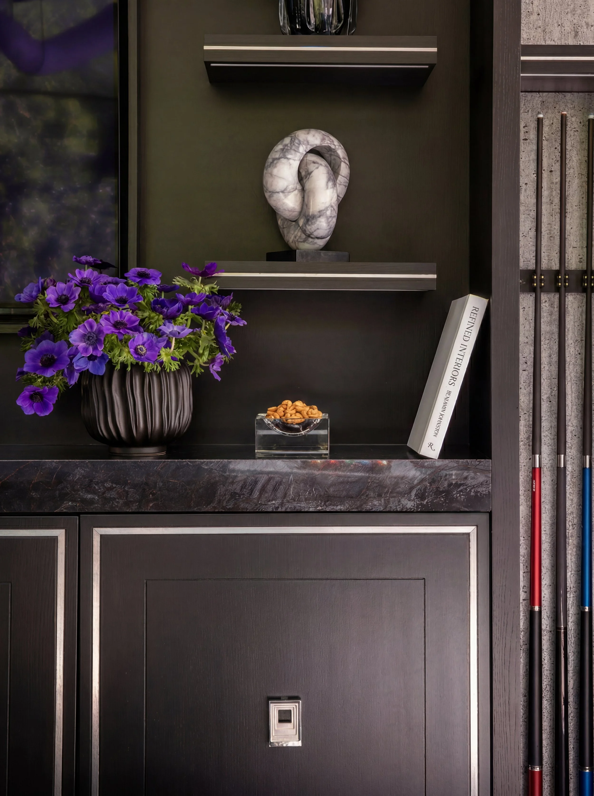 Refined Interiors Timeless Homes for Modern Living by Benjamin Johnston published by Rizzoli displayed on dark built-in shelving alongside marble sculpture, purple anemone floral arrangement, and billiard cues in a Benjamin Johnston Design game room.
