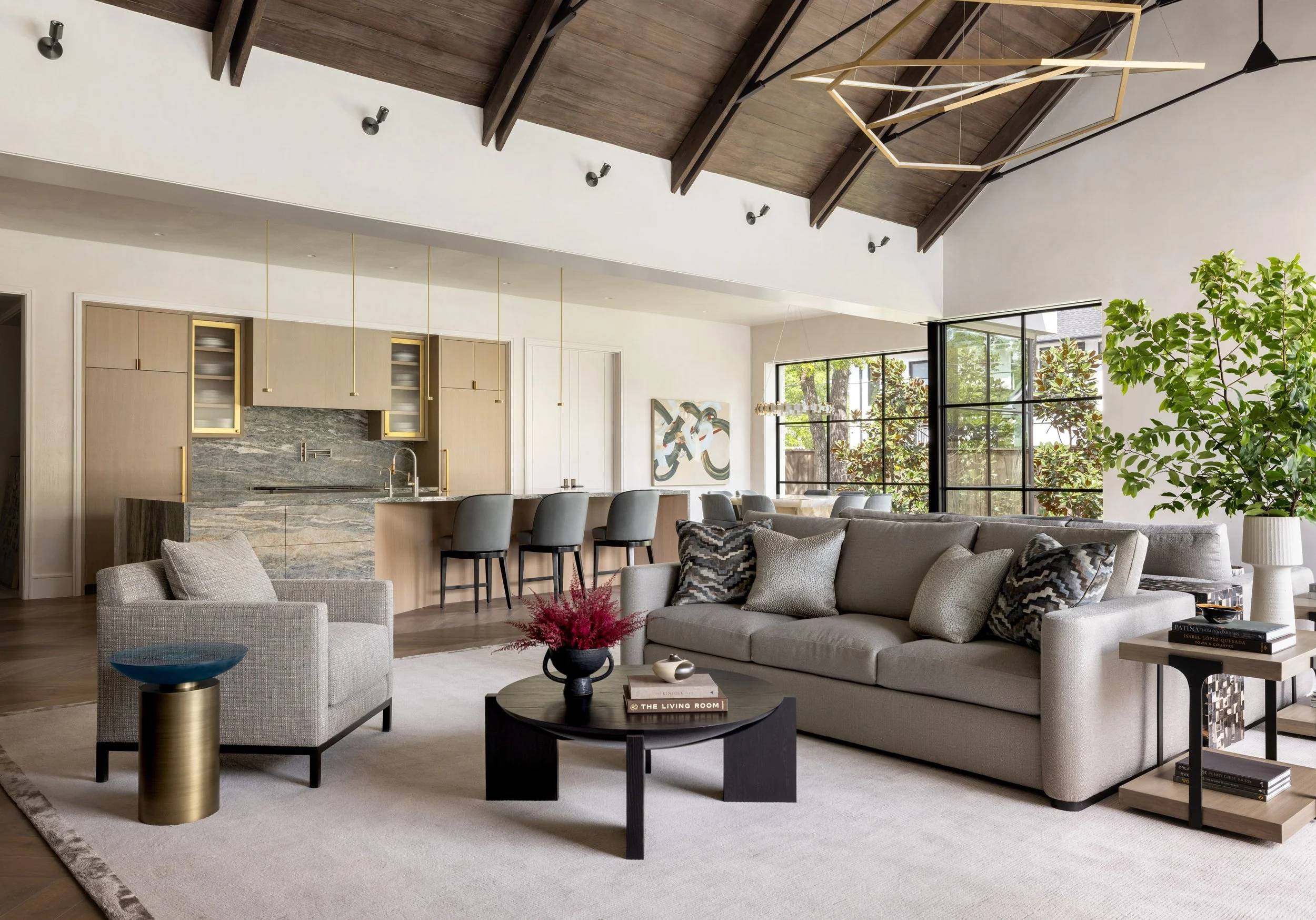 Benjamin Johnston Design open plan living and kitchen featuring vaulted wood ceiling with exposed beams, geometric brass chandelier, gray sofa with patterned pillows, sculptural black coffee table, and marble waterfall island.