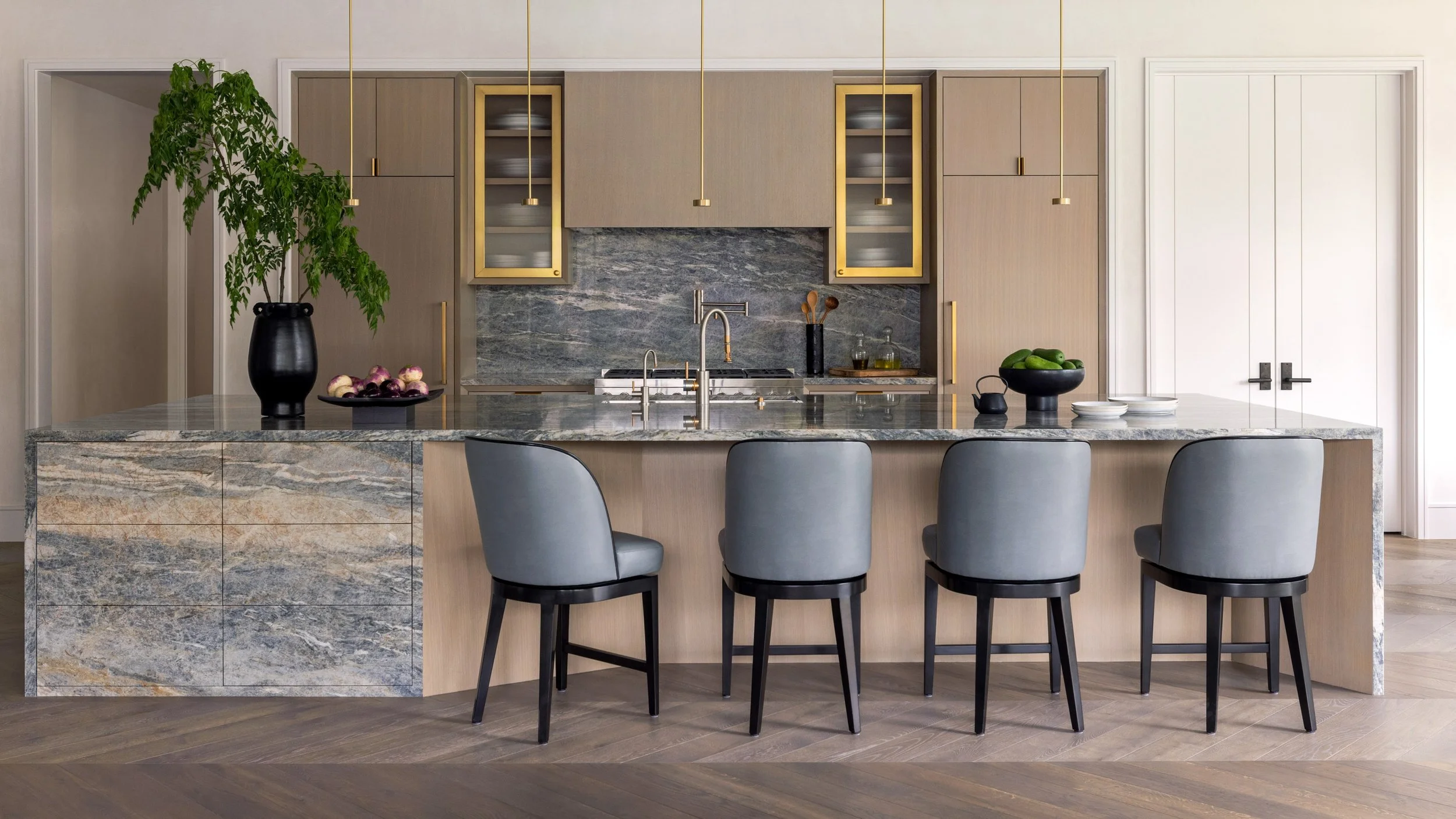 Benjamin Johnston Design kitchen with flat front cabinetry, brass framed glass uppers, veined marble backsplash, waterfall stone island, gray leather bar stools, brass pendant lights, and herringbone wood floor.
