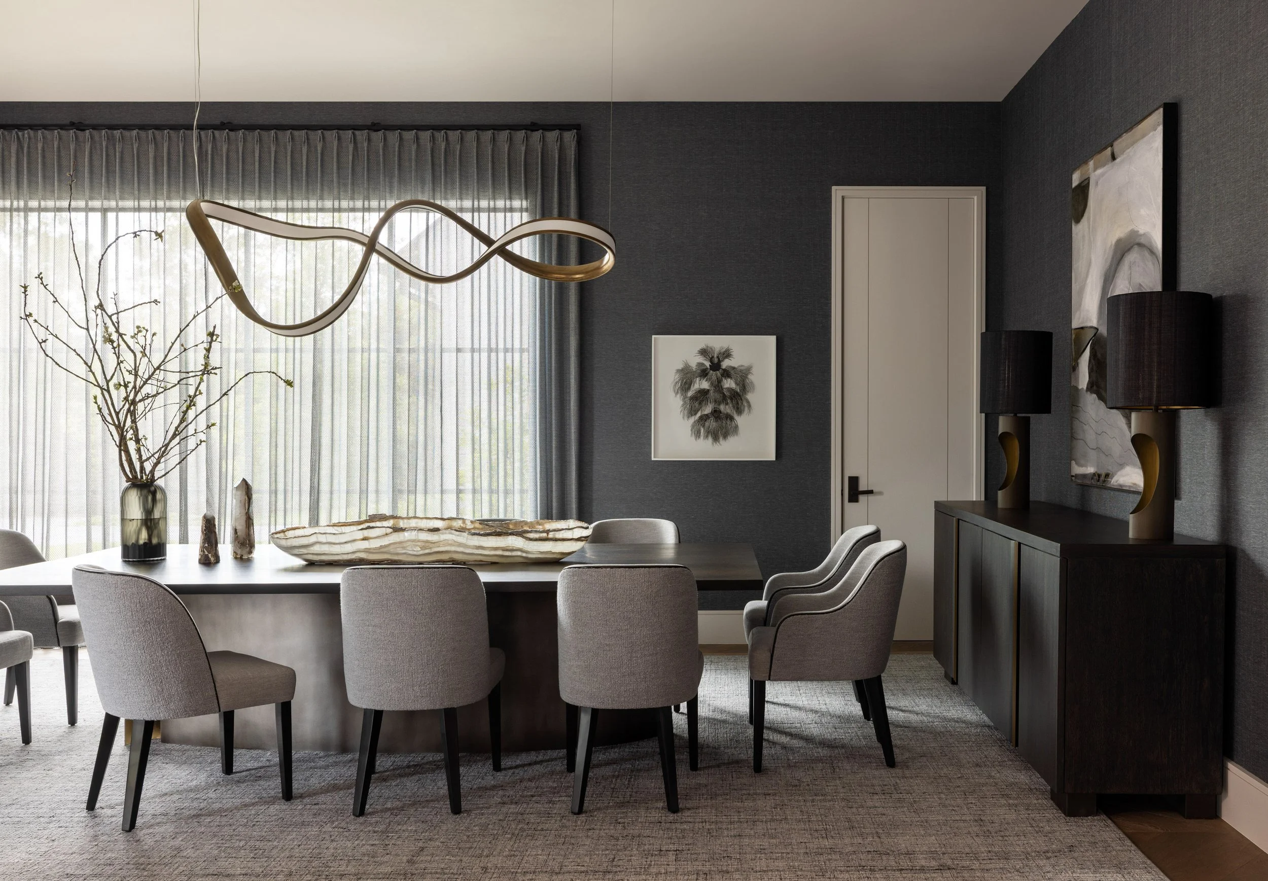 Benjamin Johnston Design Houston formal dining room featuring dark grasscloth walls, sculptural brass ribbon chandelier, gray upholstered dining chairs, petrified wood centerpiece, dark sideboard with brass lamps, and botanical artwork.
