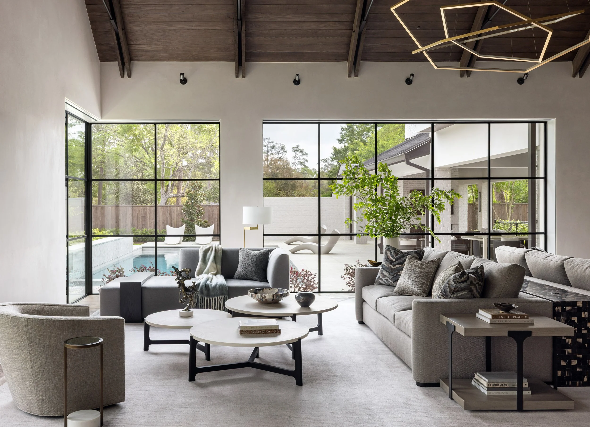 Benjamin Johnston Design indoor outdoor living room featuring expansive steel window walls opening to pool and courtyard, geometric brass chandelier, vaulted wood ceiling, gray and neutral sectional sofas, tiered round coffee tables, and sculptural.