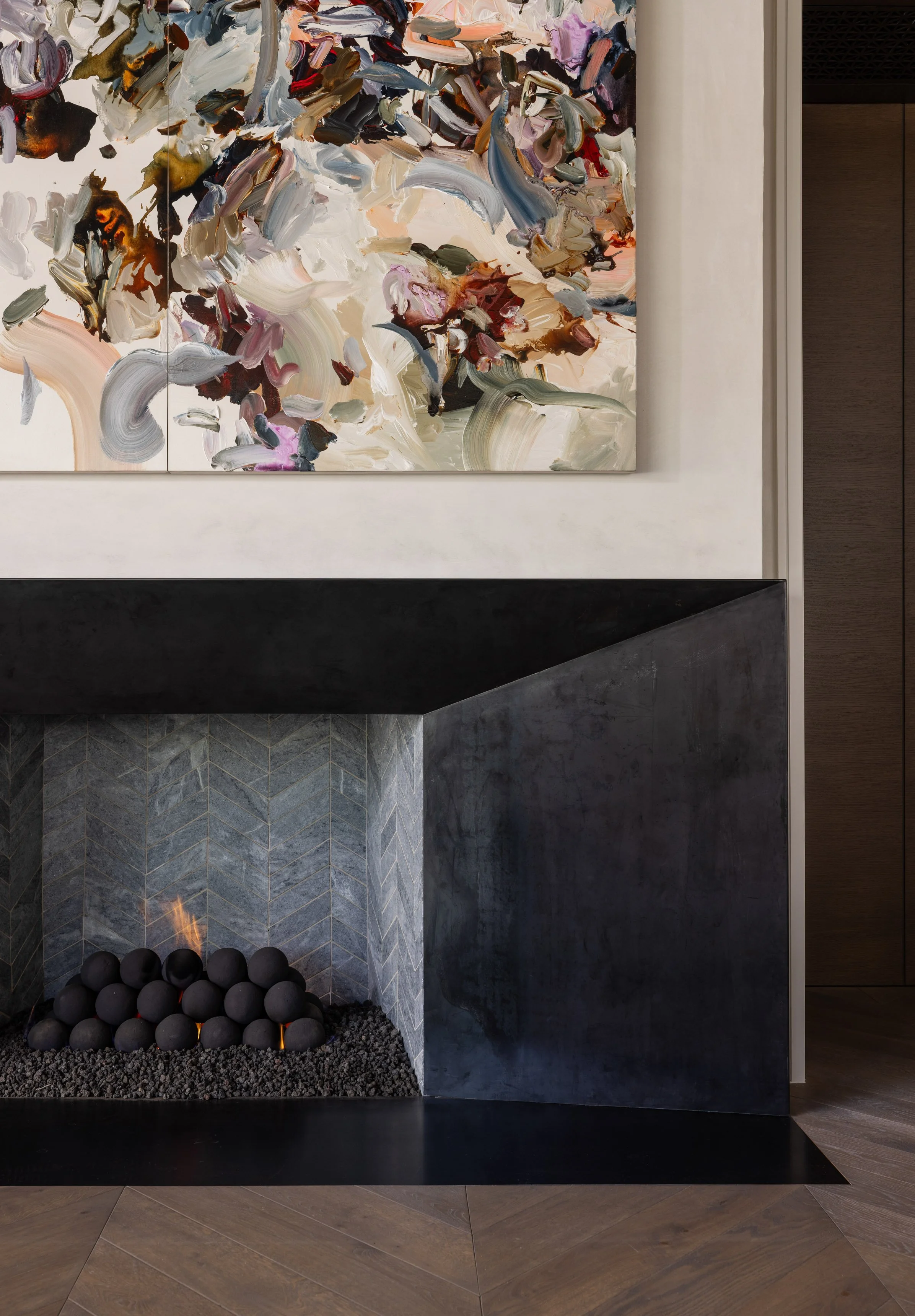 Benjamin Johnston Design fireplace close-up detail featuring sculptural dark plaster surround, chevron marble tile firebox, ceramic ball fire feature with live flame, and large scale abstract triptych artwork above.