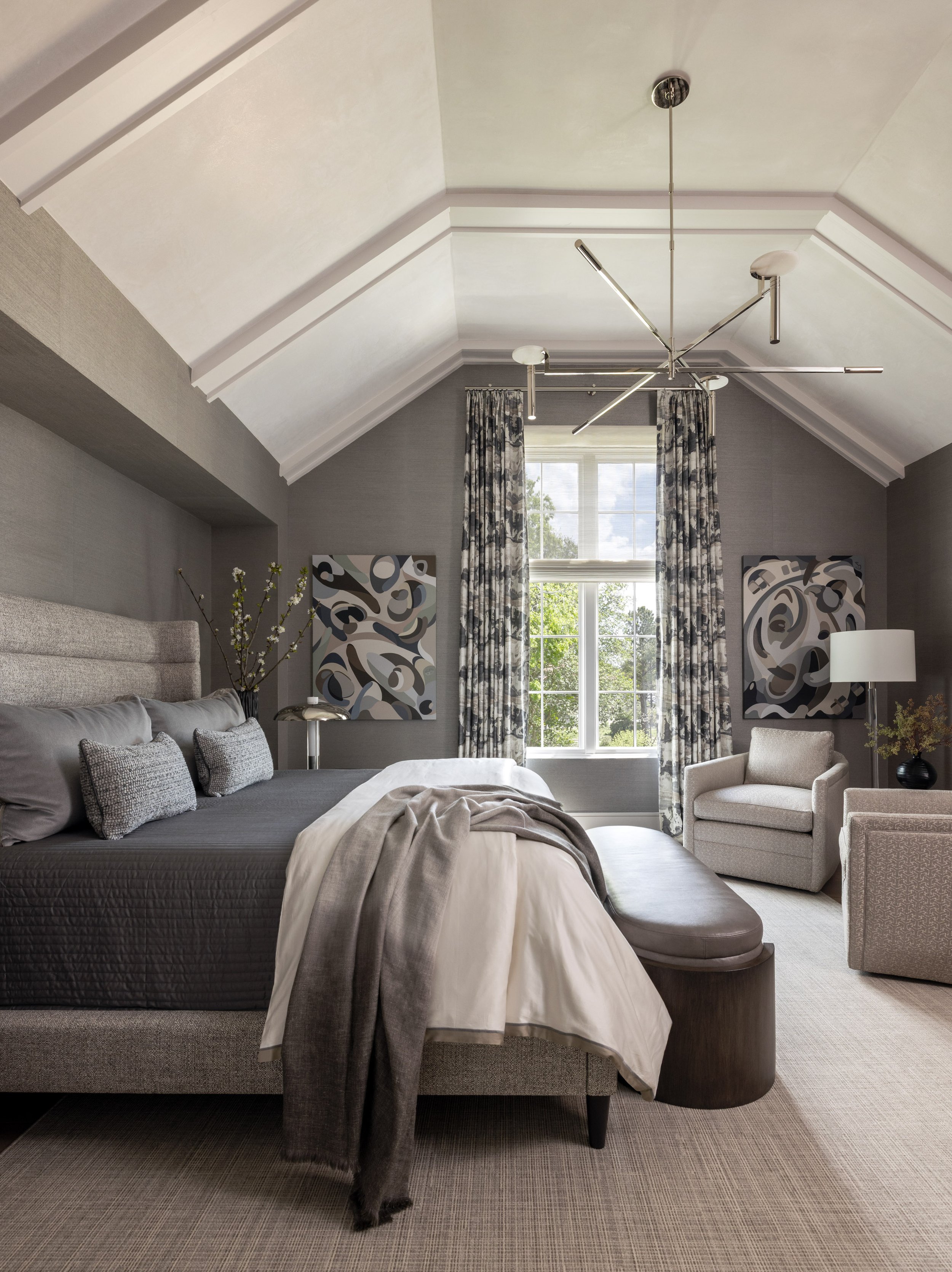 A bedroom with a sloped ceiling, a large window with curtains, abstract paintings, a bed with gray and white bedding, a gray upholstered headboard, a round ottoman at the foot of the bed, and a black nightstand with a decorative plant.