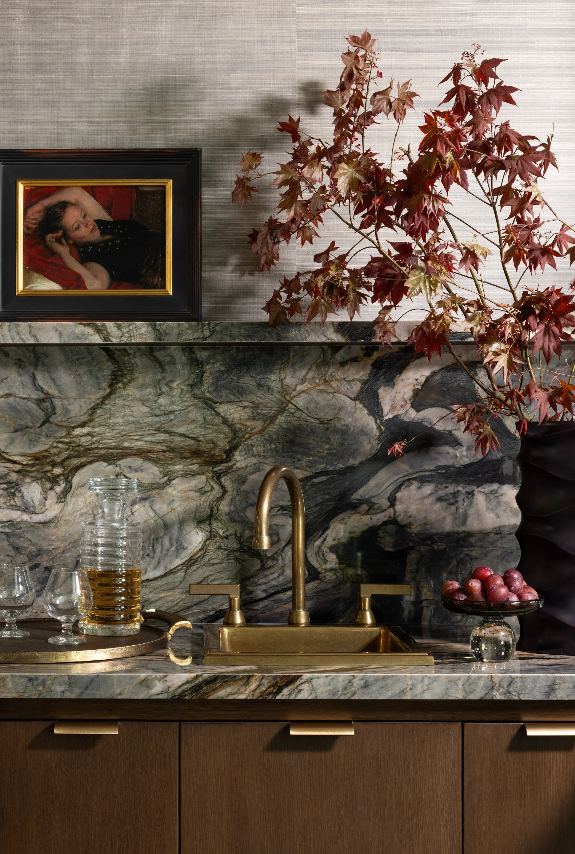 Benjamin Johnston Design wet bar detail with dramatic veined marble backsplash, brass sink and faucet, brass serving tray with crystal decanter, red maple branches, framed figurative painting, and grasscloth wallcovering.