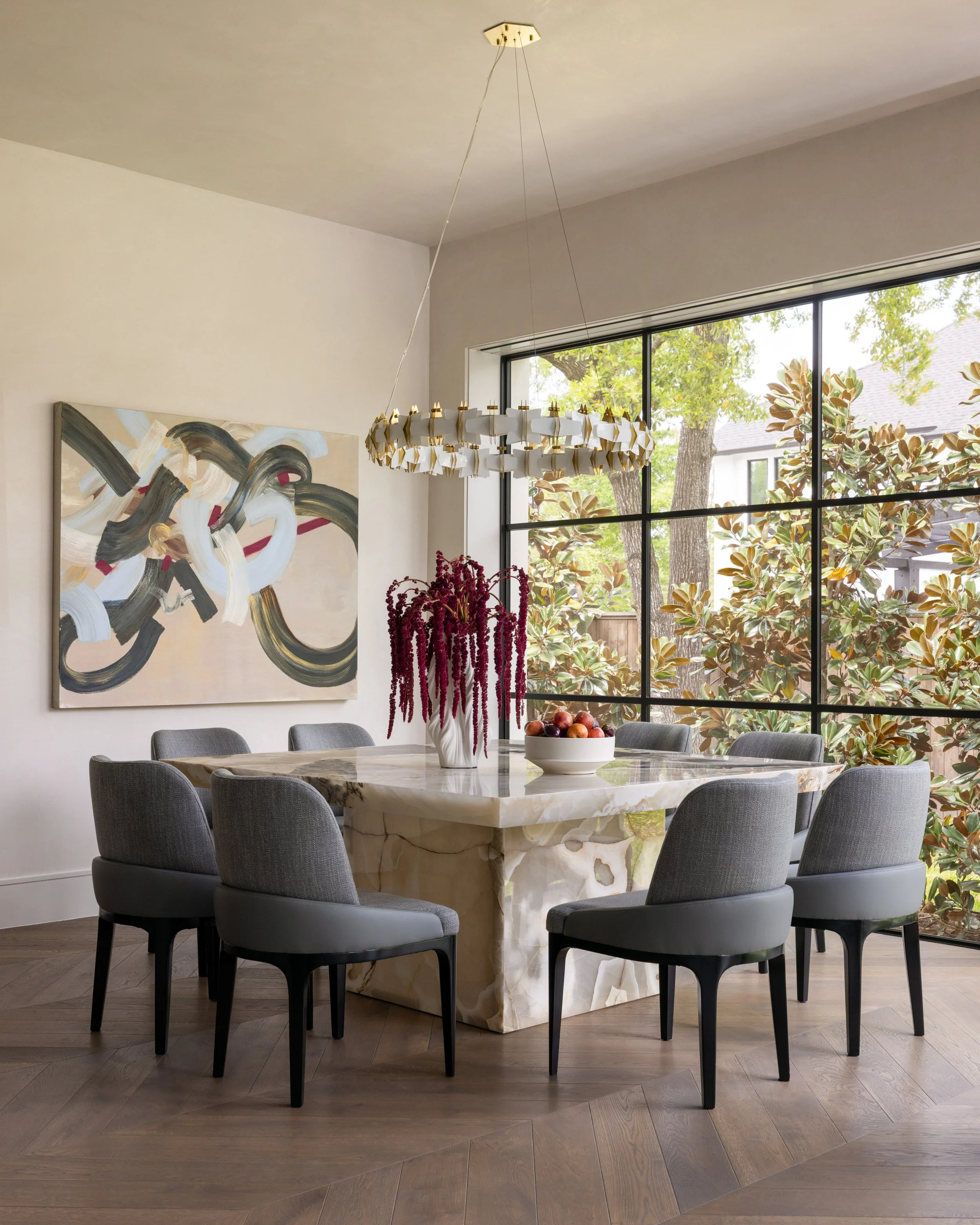 A modern dining room with a large marble table, six gray upholstered chairs, a white and gold chandelier, a fruit bowl, a flower arrangement, an abstract painting on the wall, and large window with views of trees and a neighboring house.