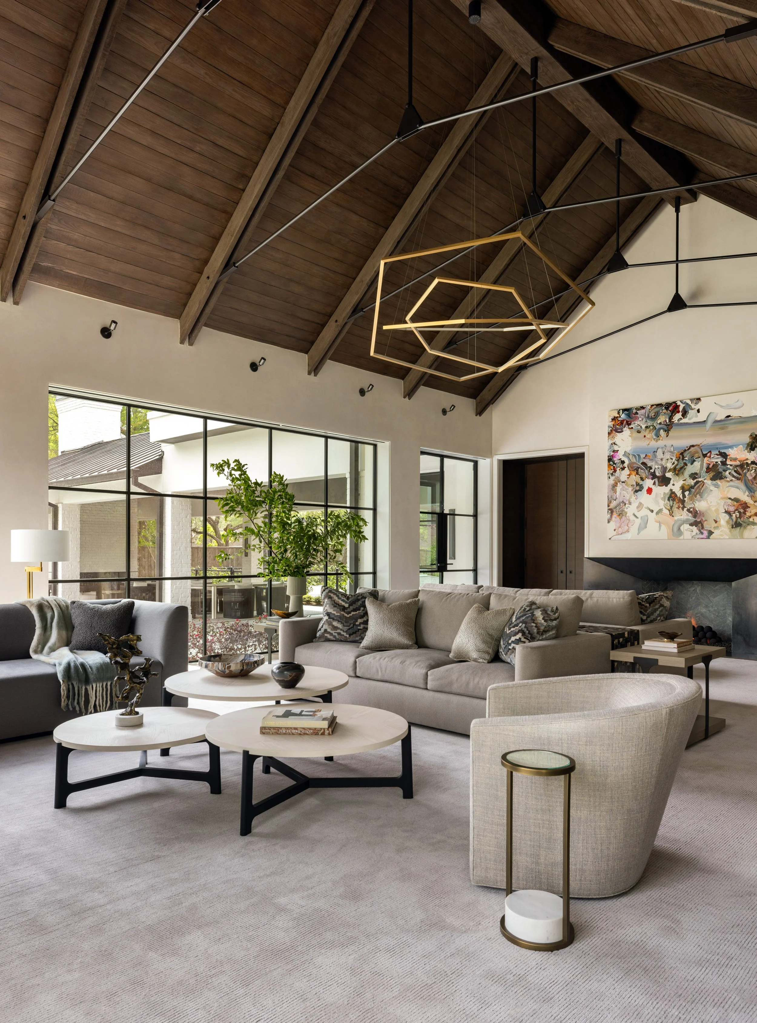 Benjamin Johnston Design living room detail featuring geometric brass chandelier, vaulted wood ceiling with exposed beams, curved lounge chair, tiered round coffee tables, neutral sectional sofa with patterned pillows, and stone fireplace.