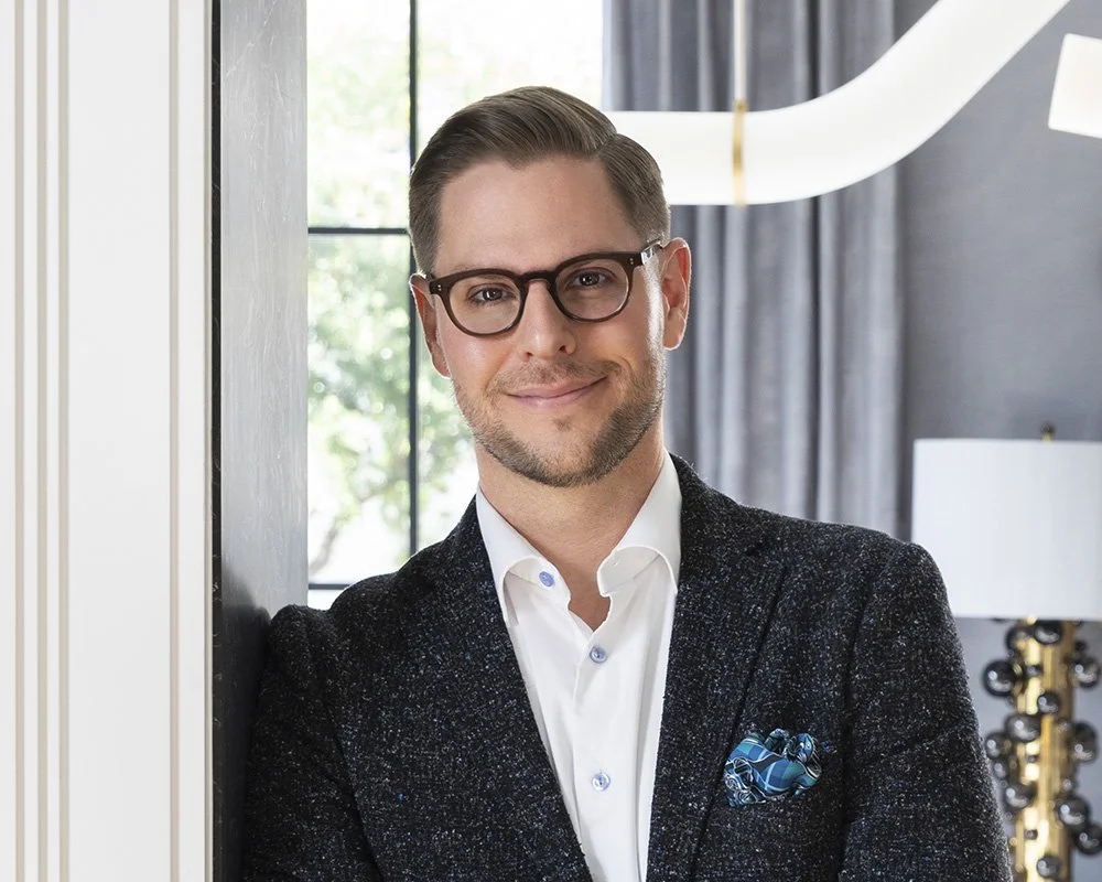 A man with short brown hair, glasses, and a beard, smiling and wearing a dark blazer over a white shirt with a blue pocket square, standing indoors against a wall with a large window in the background.