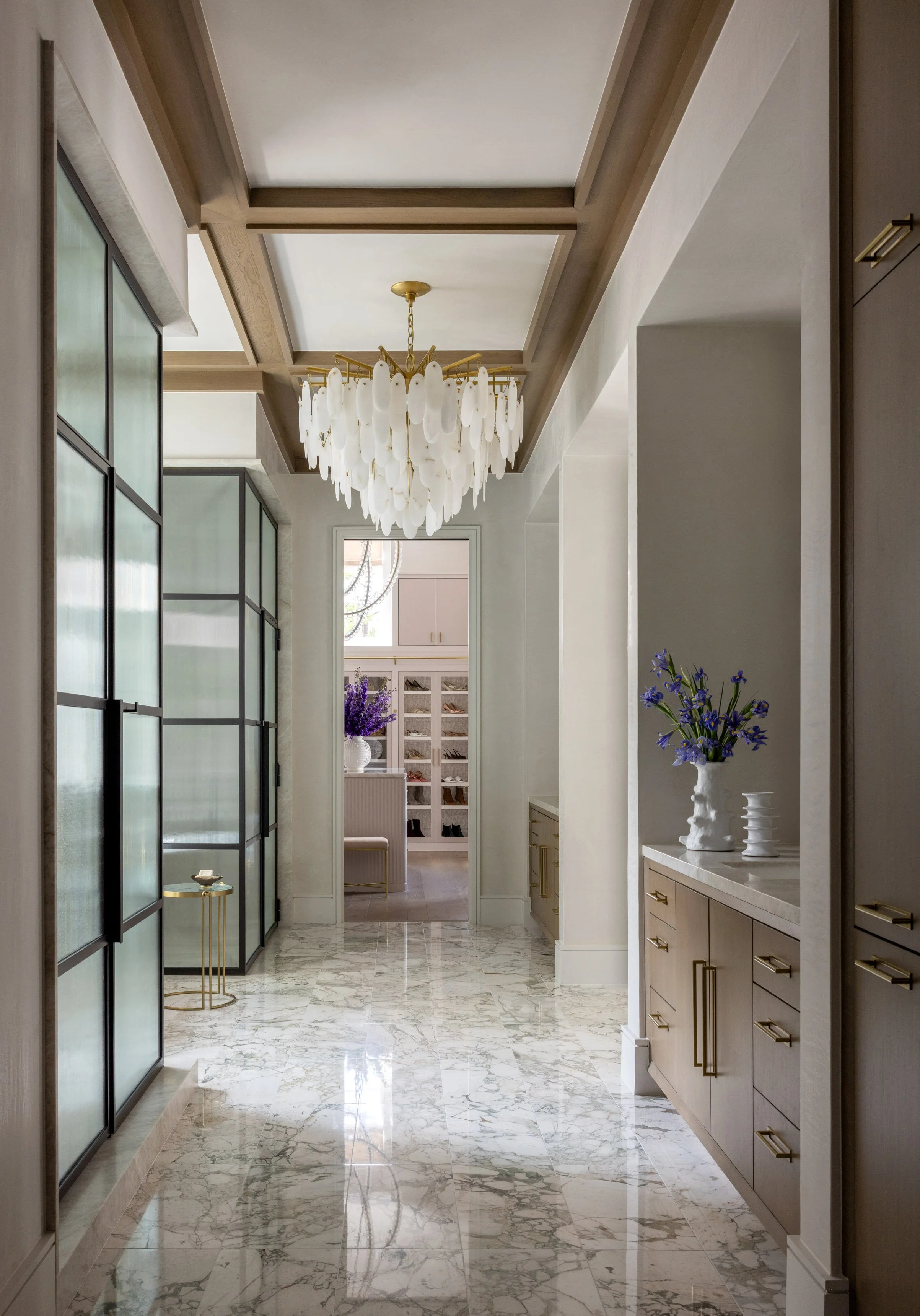 A hallway with marble flooring, beige cabinetry, and a decorative chandelier hanging from the ceiling. A vase with purple flowers is on the right side, and a small side table is on the left. Doors and windows are visible in the background.