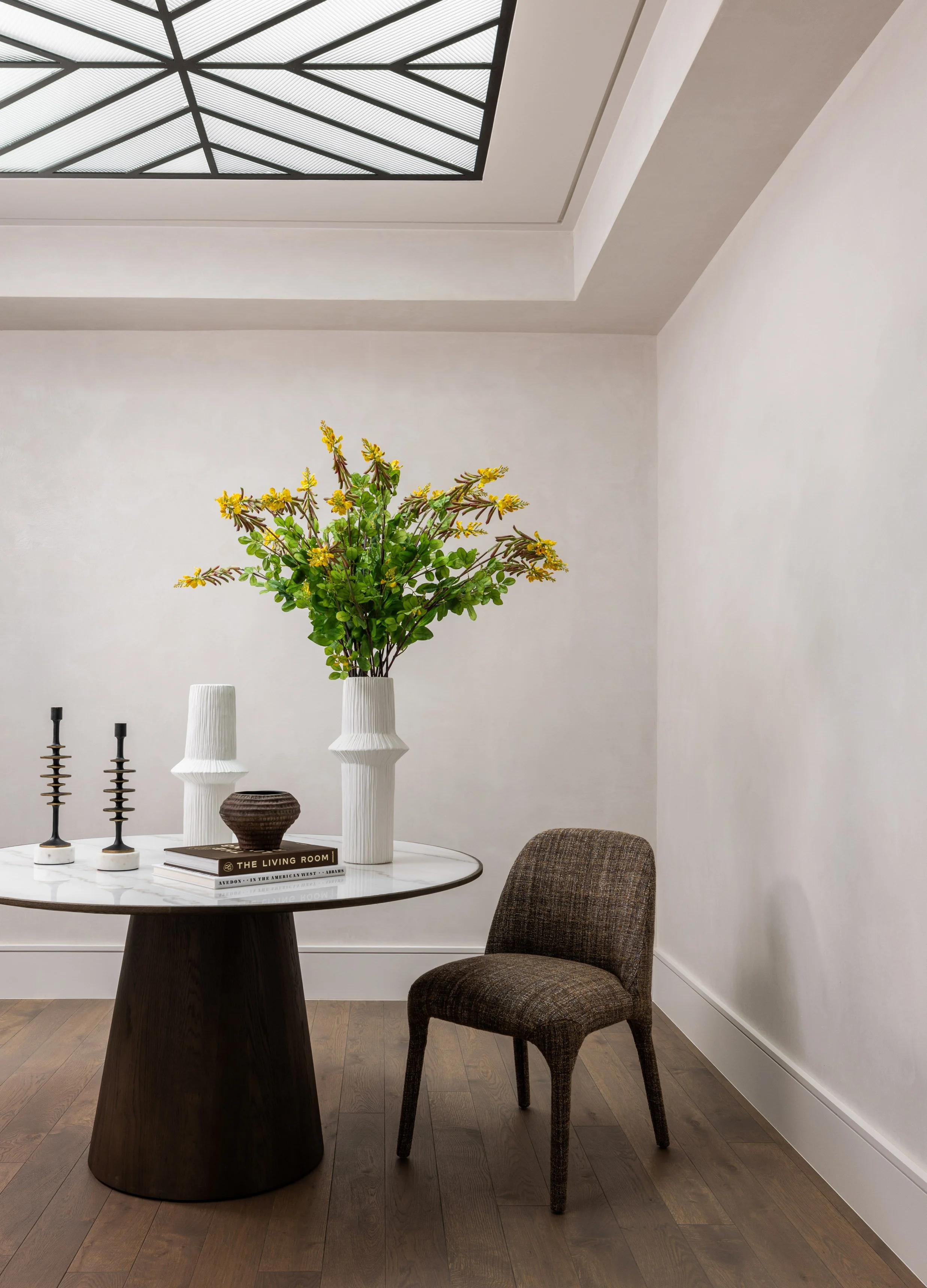 A round dining table with a black base and a white marble top, decorated with a black vase with yellow flowers, a stack of books, and two brass candlestick holders. There is a brown upholstered chair beside the table. The background features plain wh