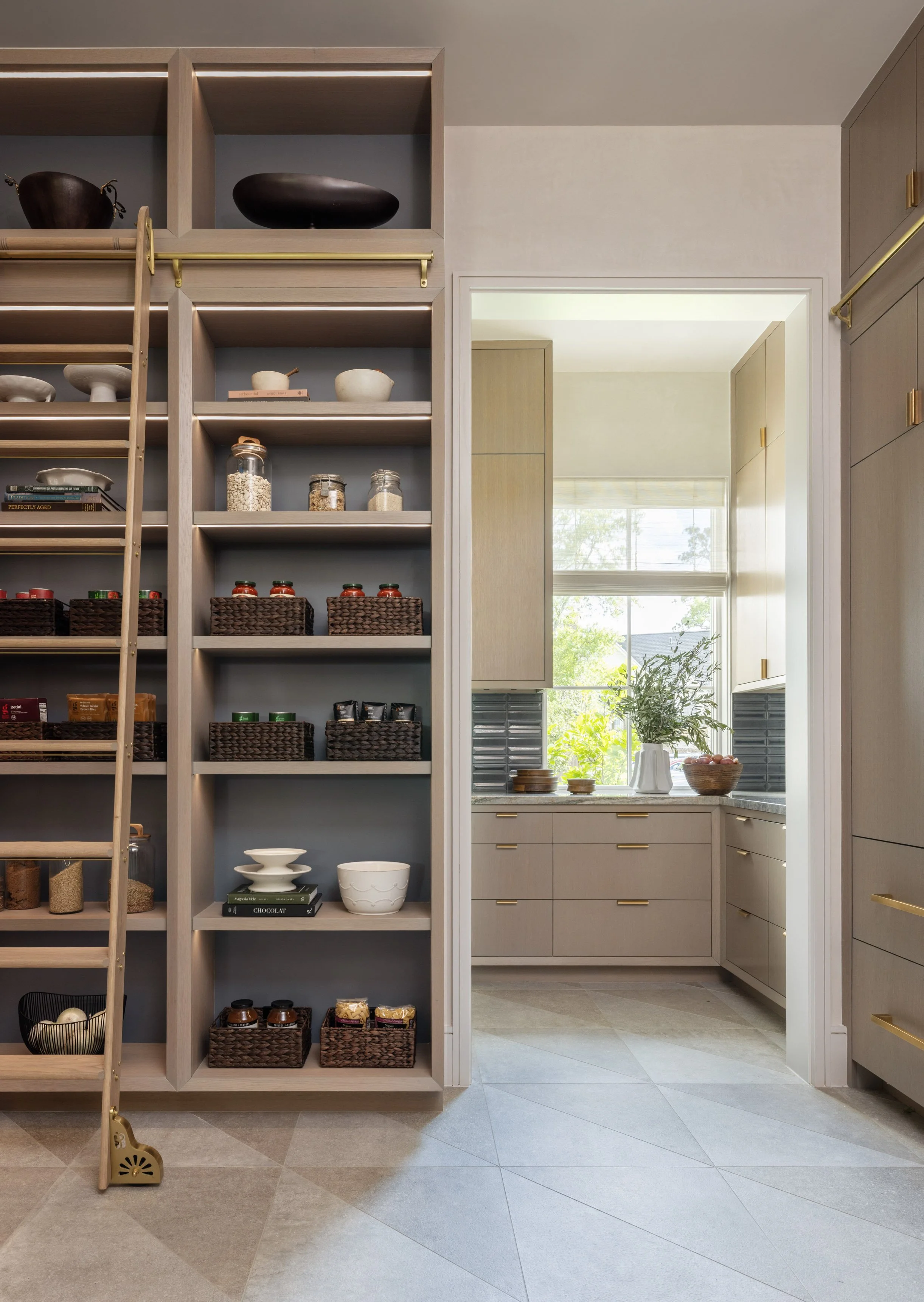Benjamin Johnston Design Houston kitchen pantry featuring floor-to-ceiling oak shelving with brass library ladder, organized woven baskets, and view into catering kitchen with taupe cabinetry and brass hardware.