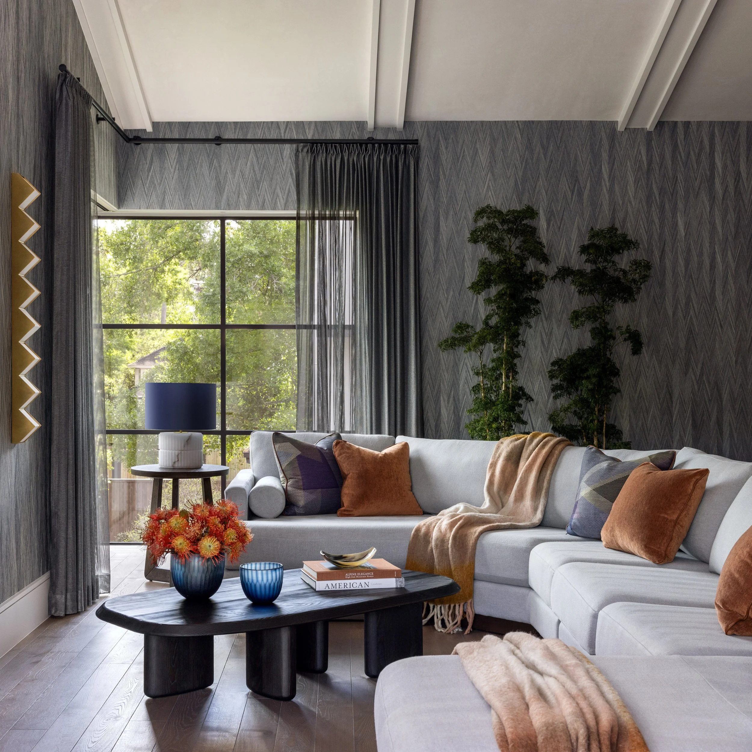 Living room with gray chevron-patterned walls, large window with black sheer curtains showing greenery outside, white sofa with deep orange and striped pillows, dark wooden coffee table with a bouquet of orange flowers, books, and blue vases, and a b