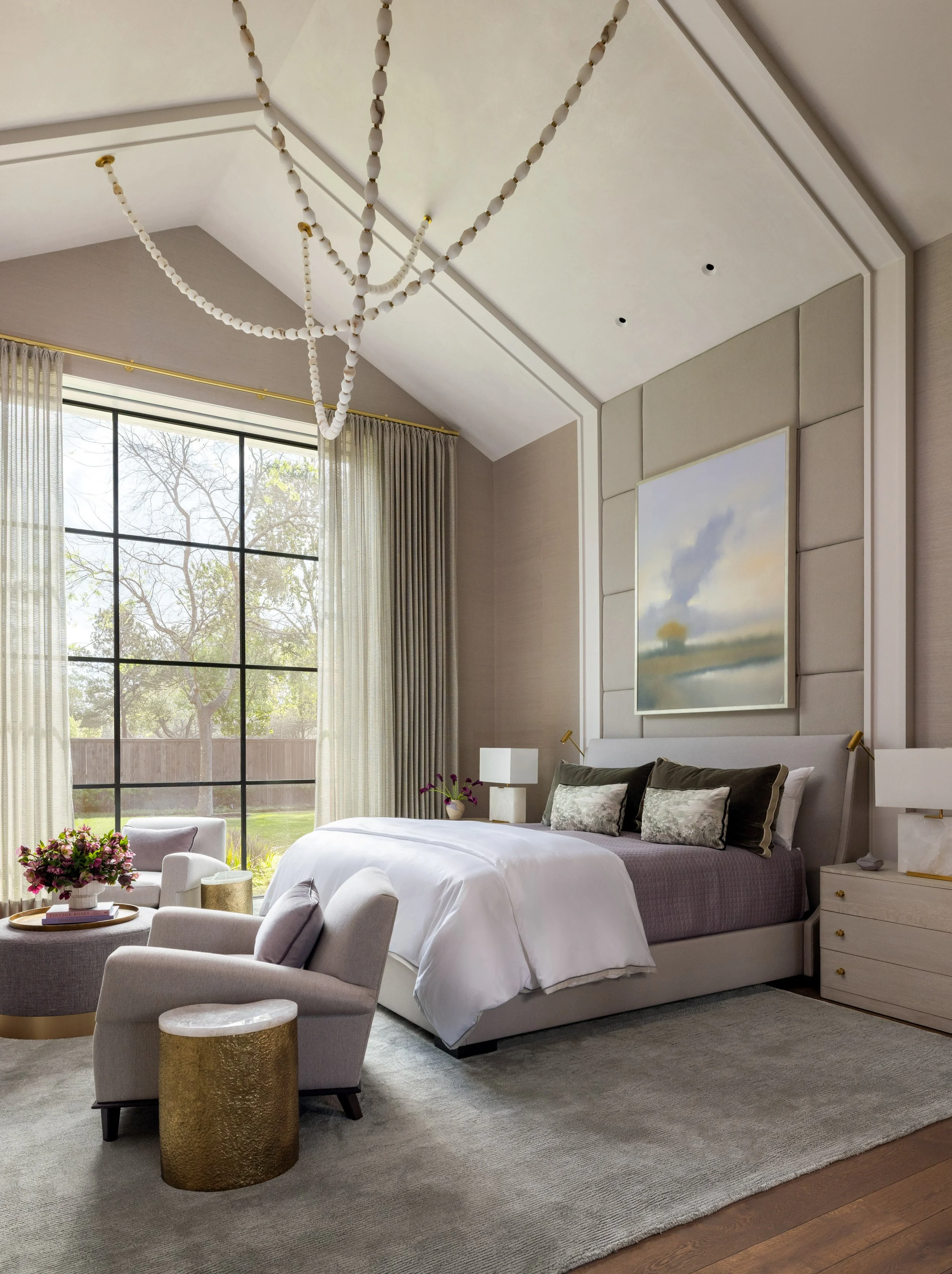 Benjamin Johnston Design primary bedroom wide view featuring sculptural beaded chandelier, vaulted ceiling with architectural molding, upholstered panel headboard wall, soft landscape artwork, sitting area with lavender lounge chairs, and brass.