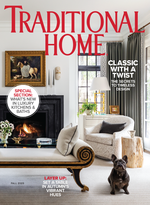 A cozy living room with a fireplace, black mantel, and a framed painting above it, decorated with candles and flowers. There is a cream-colored chaise lounge with pillows, a white armchair, a wooden side table, and a small dog sitting on the floor. Large windows with dark curtains let in natural light.
