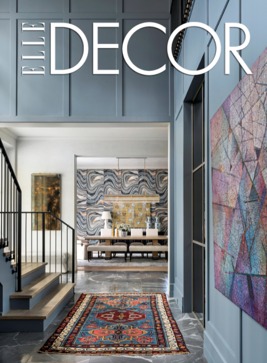 Interior view of an upscale home with blue-gray walls, a colorful area rug, and modern artwork, featuring a staircase with black railing leading to a dining area with abstract wall art.