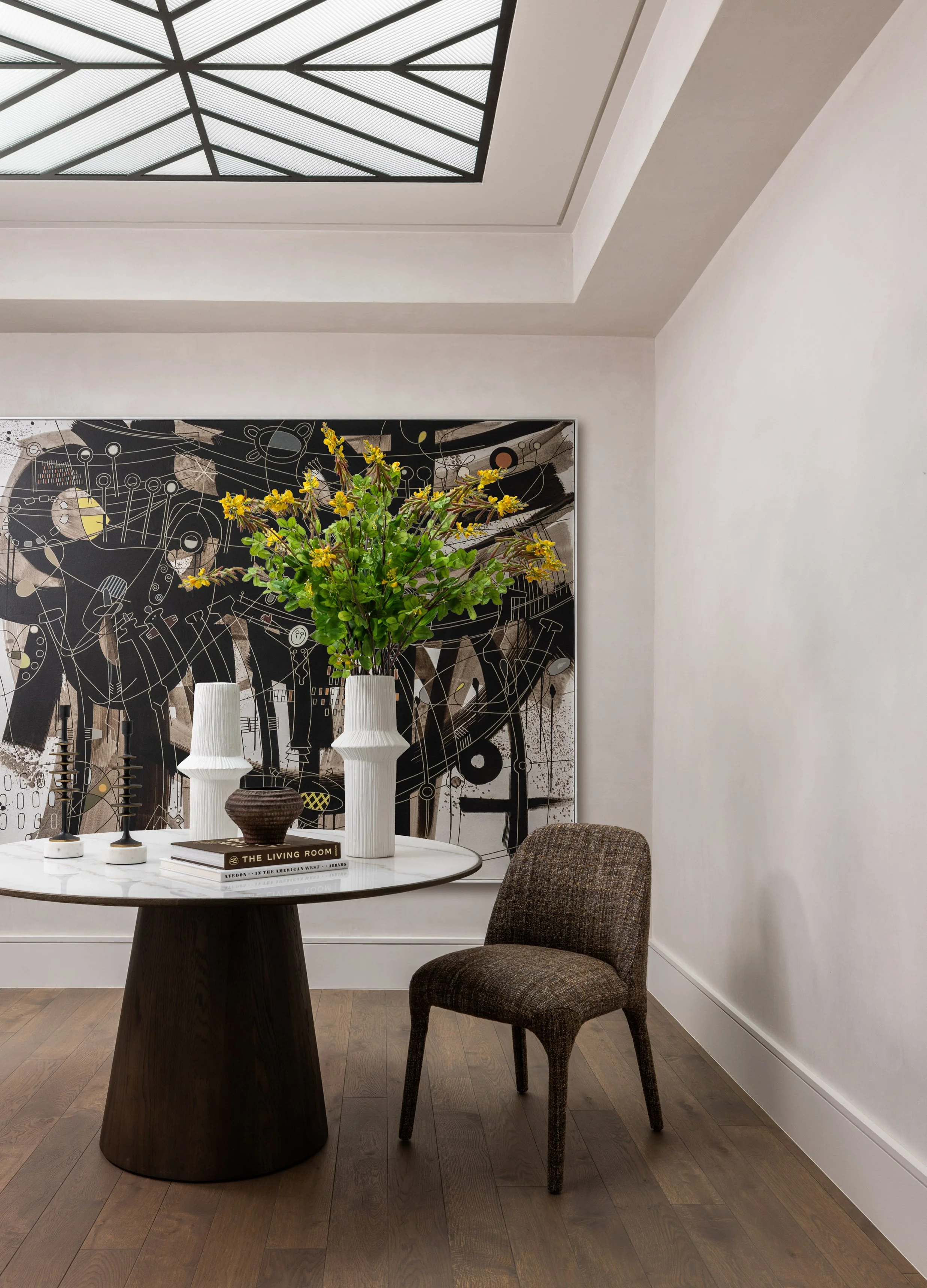 Benjamin Johnston Design entryway featuring steel and glass skylight, dark pedestal table with marble top, large scale black and white abstract artwork, textured upholstered chair, and botanical arrangement.