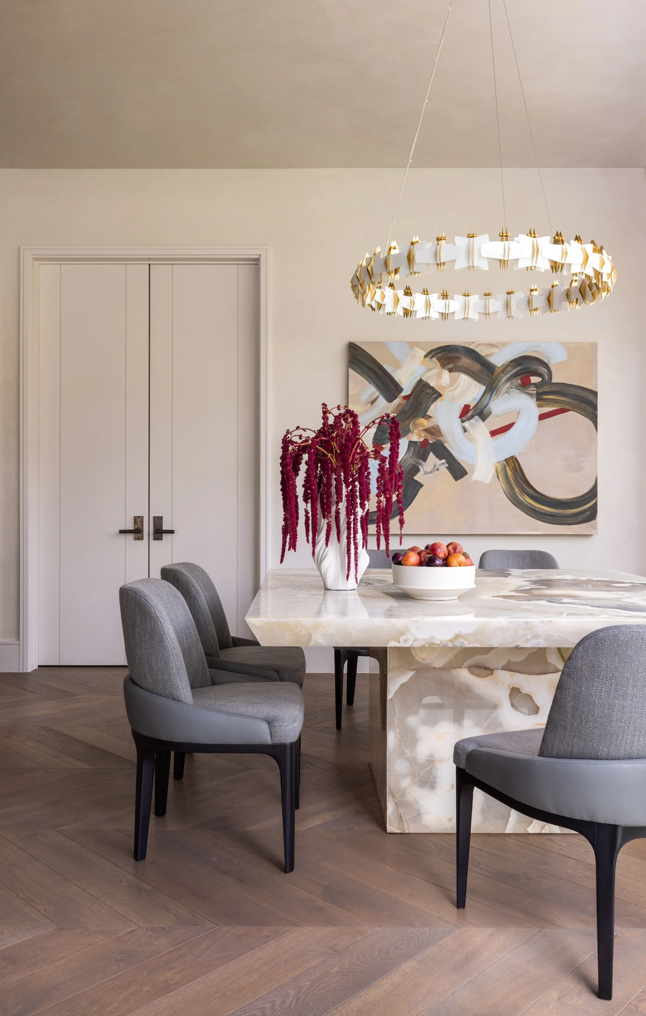 A modern dining room with a marble table, surrounded by four gray upholstered chairs. The table has a white bowl of apples and a tall white vase with red hanging amaranthus flowers. A large abstract painting and a gold and white circular chandelier a